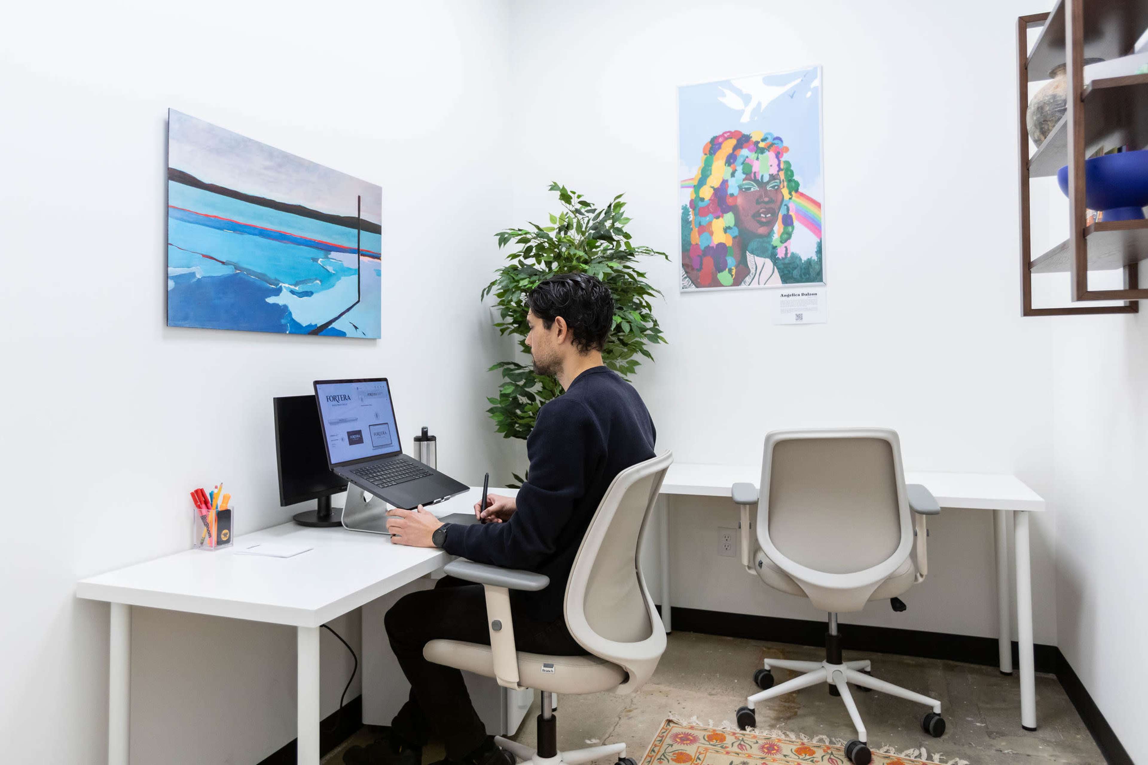 A person is working at a desk in a small office space, surrounded by artwork and a potted plant.
