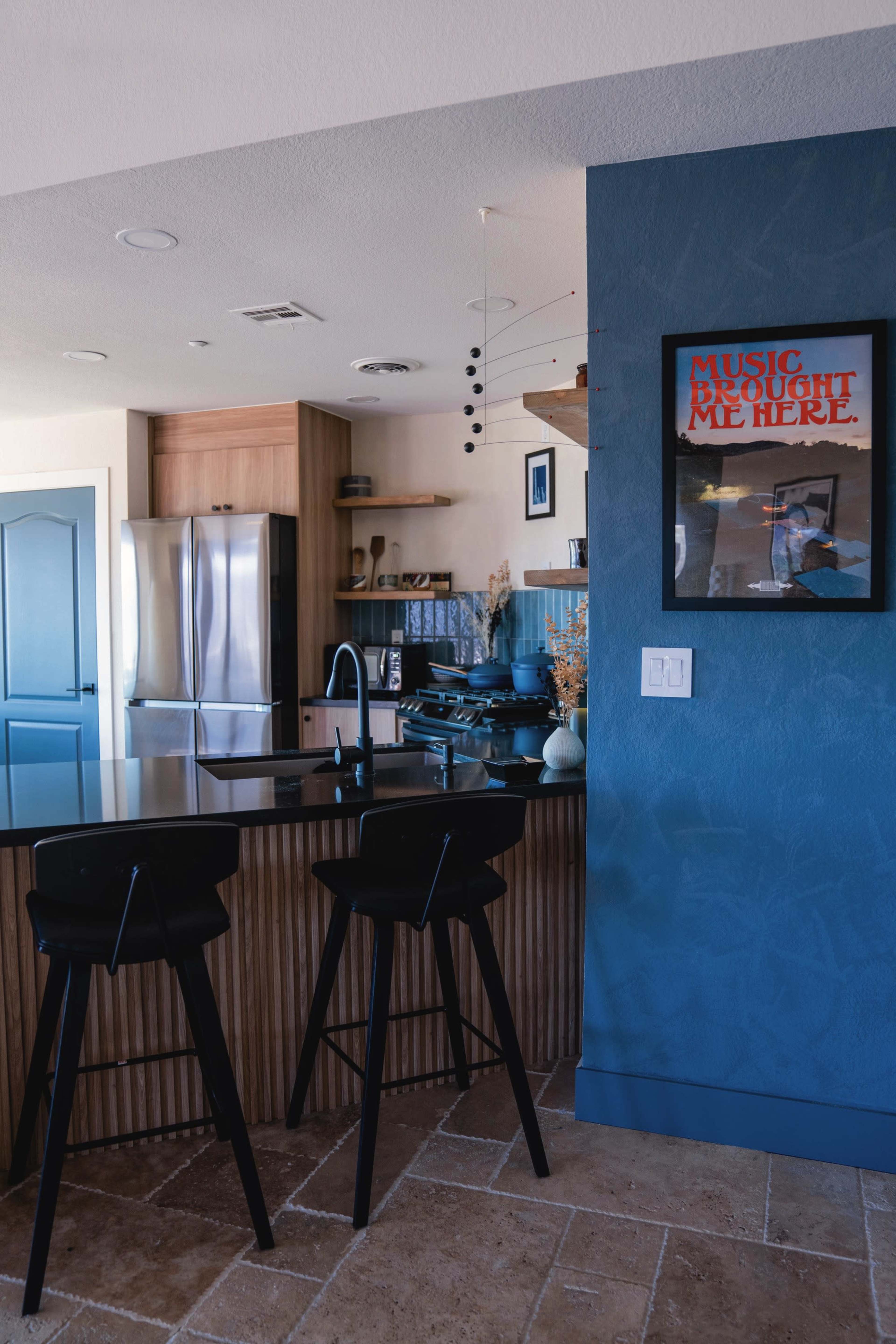 A modern kitchen features stainless steel appliances, wooden cabinetry, and a framed poster on the wall above a black island with two stools.