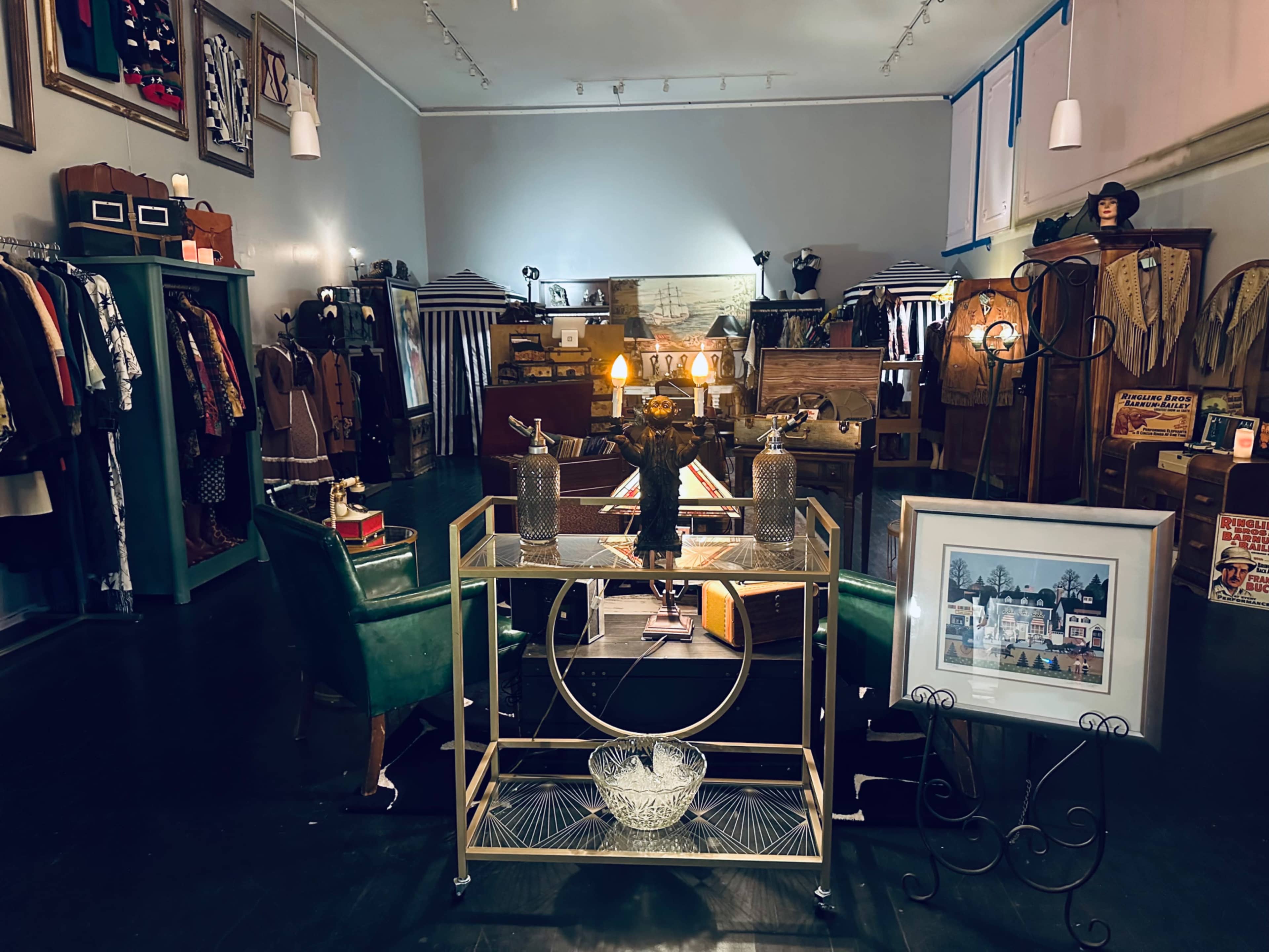 The image depicts a vintage boutique showcasing clothing racks, a seating area with green chairs, and various decor items against a neutral-colored wall.