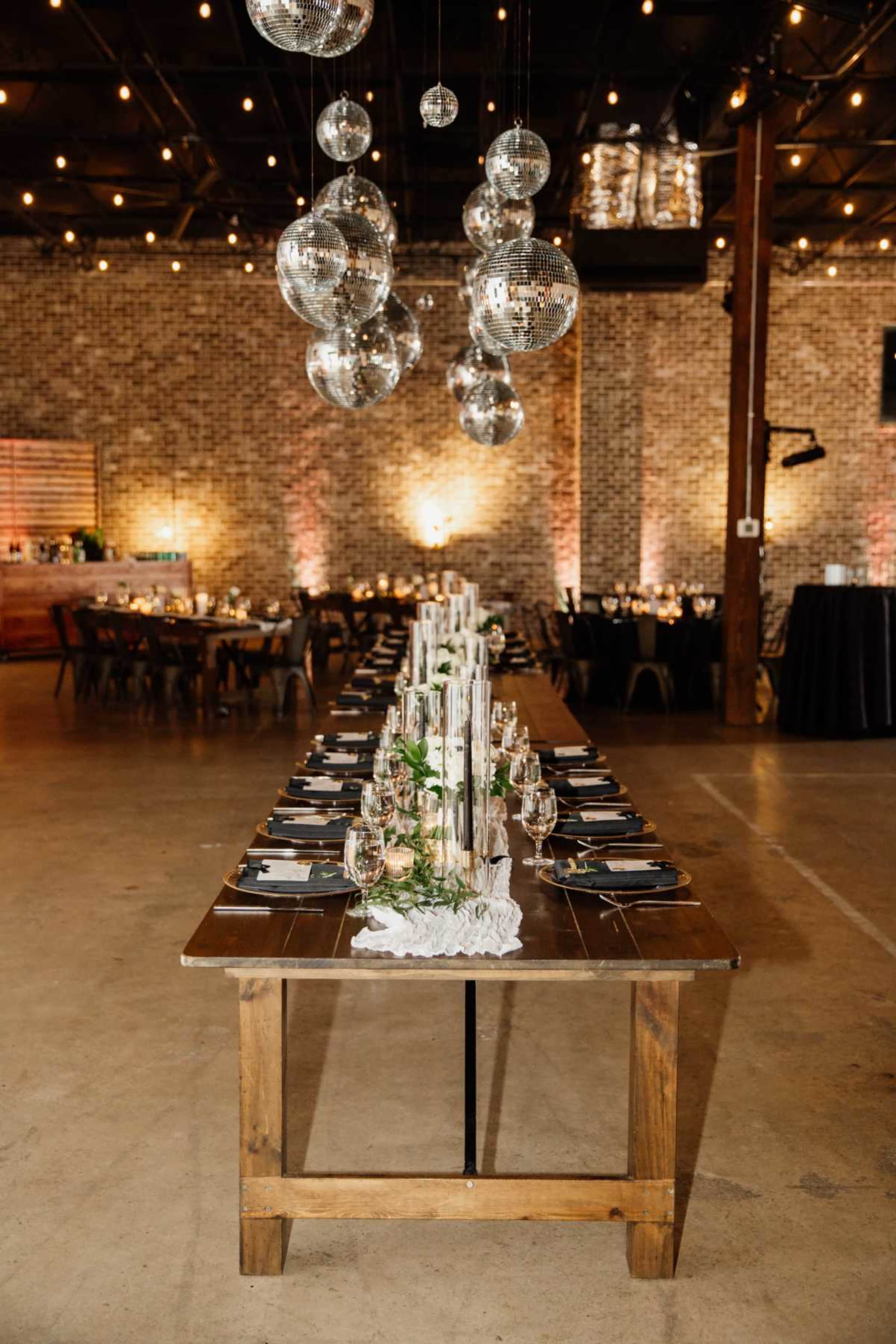 A long wooden dining table is set with plates and glasses, surrounded by string lights and hanging disco balls in a spacious venue.