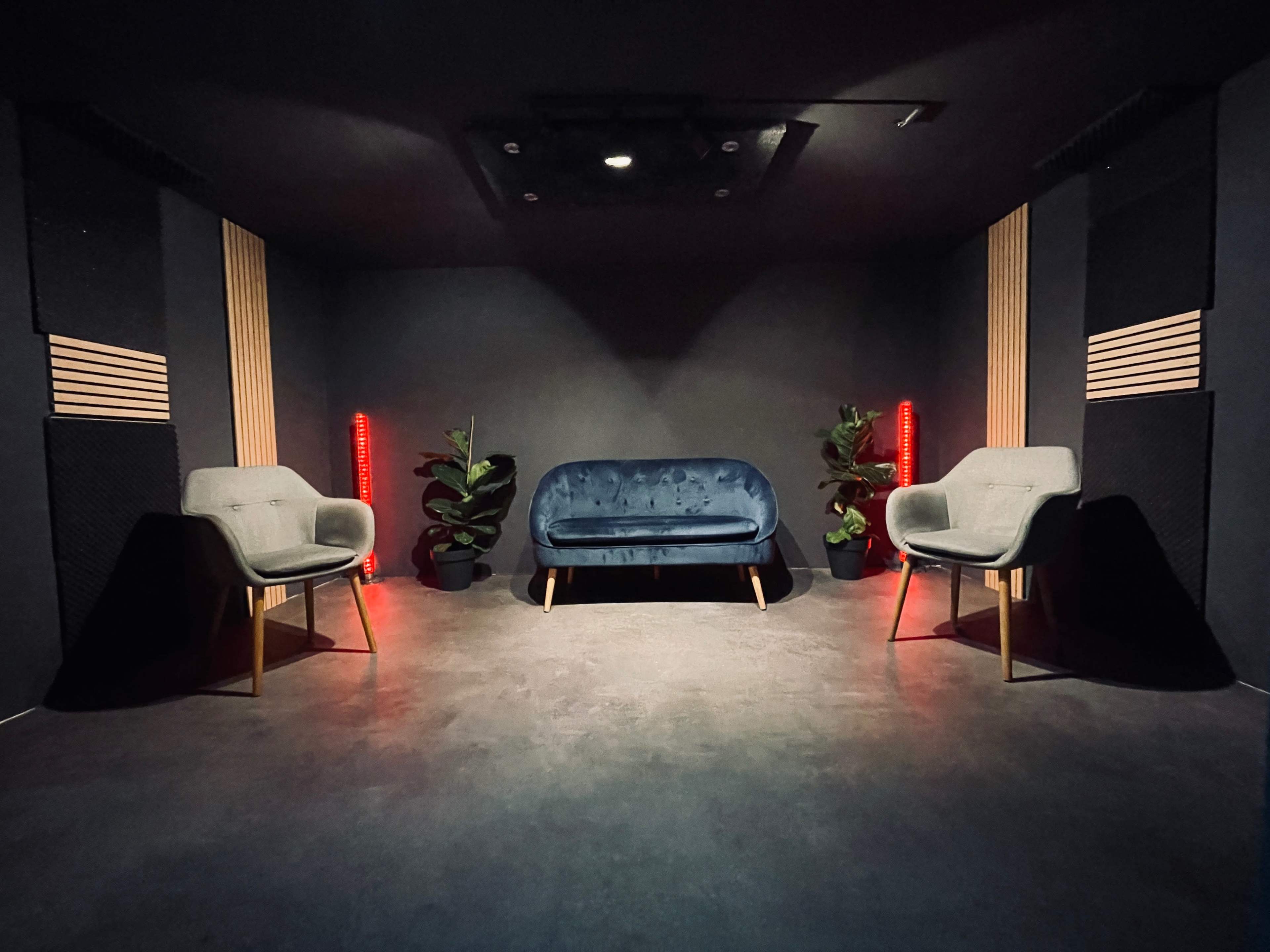 A dimly lit room featuring a blue sofa flanked by two gray chairs, with red lights and potted plants in the corners.