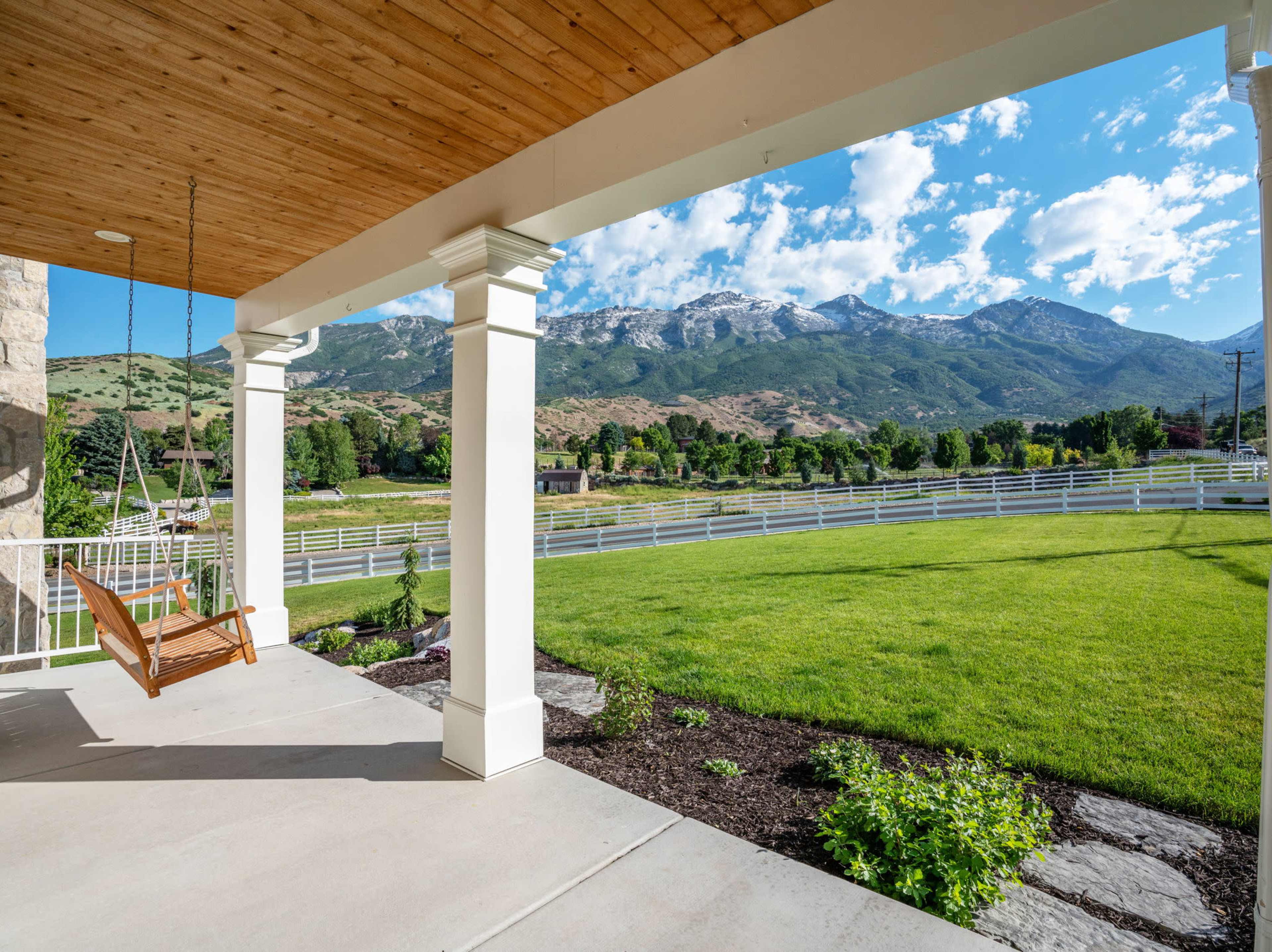A wide porch with a swing overlooks a grassy yard and mountain landscape under a partially cloudy sky.