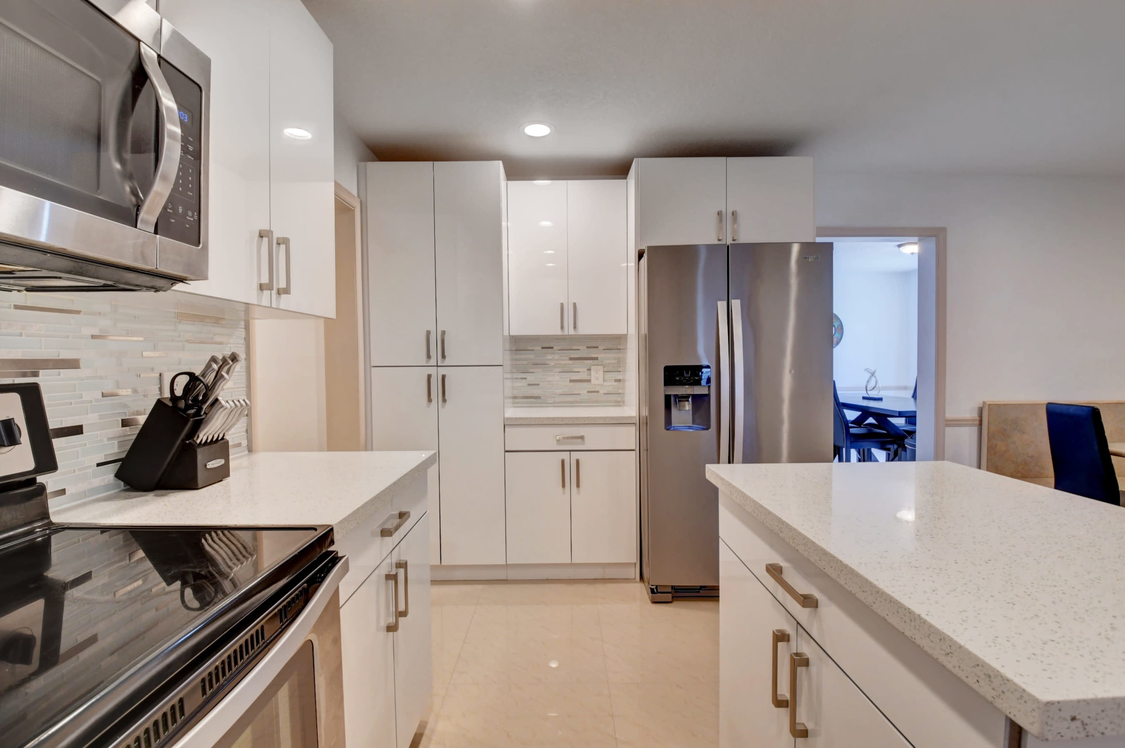 The image shows a modern kitchen with white cabinetry, a stainless steel refrigerator, and a countertop with a built-in stove.