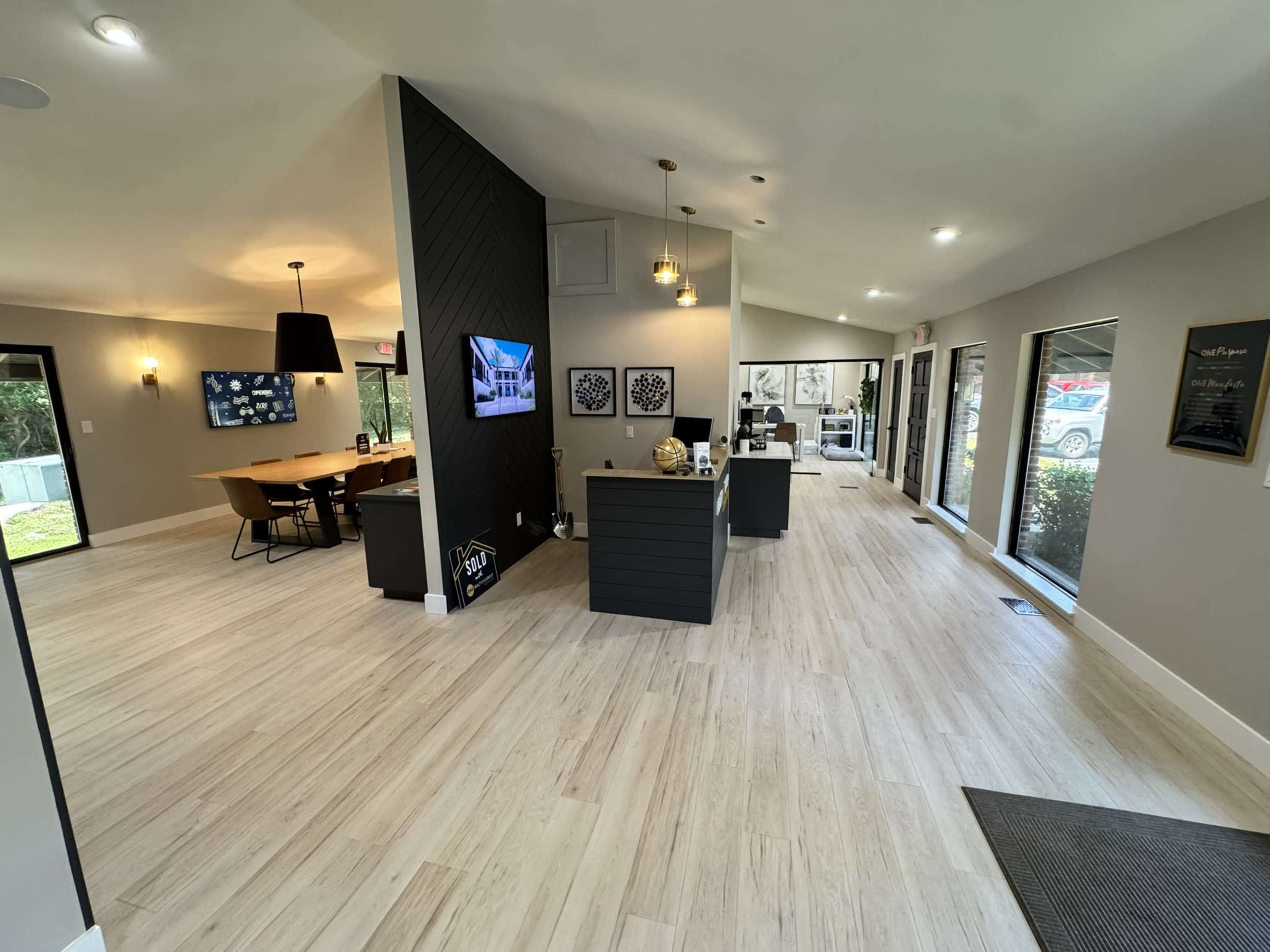 The image shows a modern lobby area with a reception desk, a dining table, a television display, and large windows providing natural light.