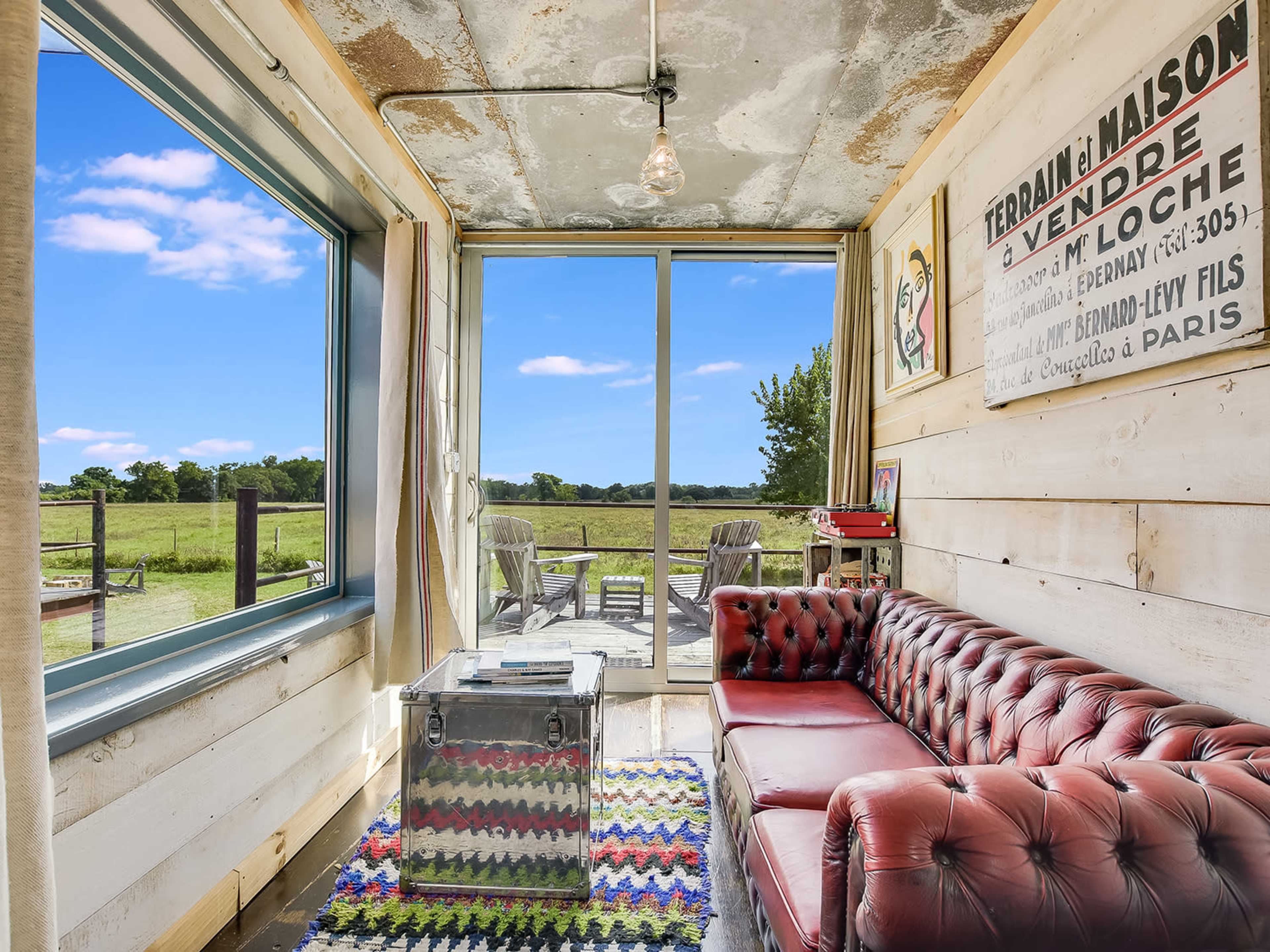A cozy room features a red leather sofa, a glass coffee table, and a large window overlooking a grassy field.