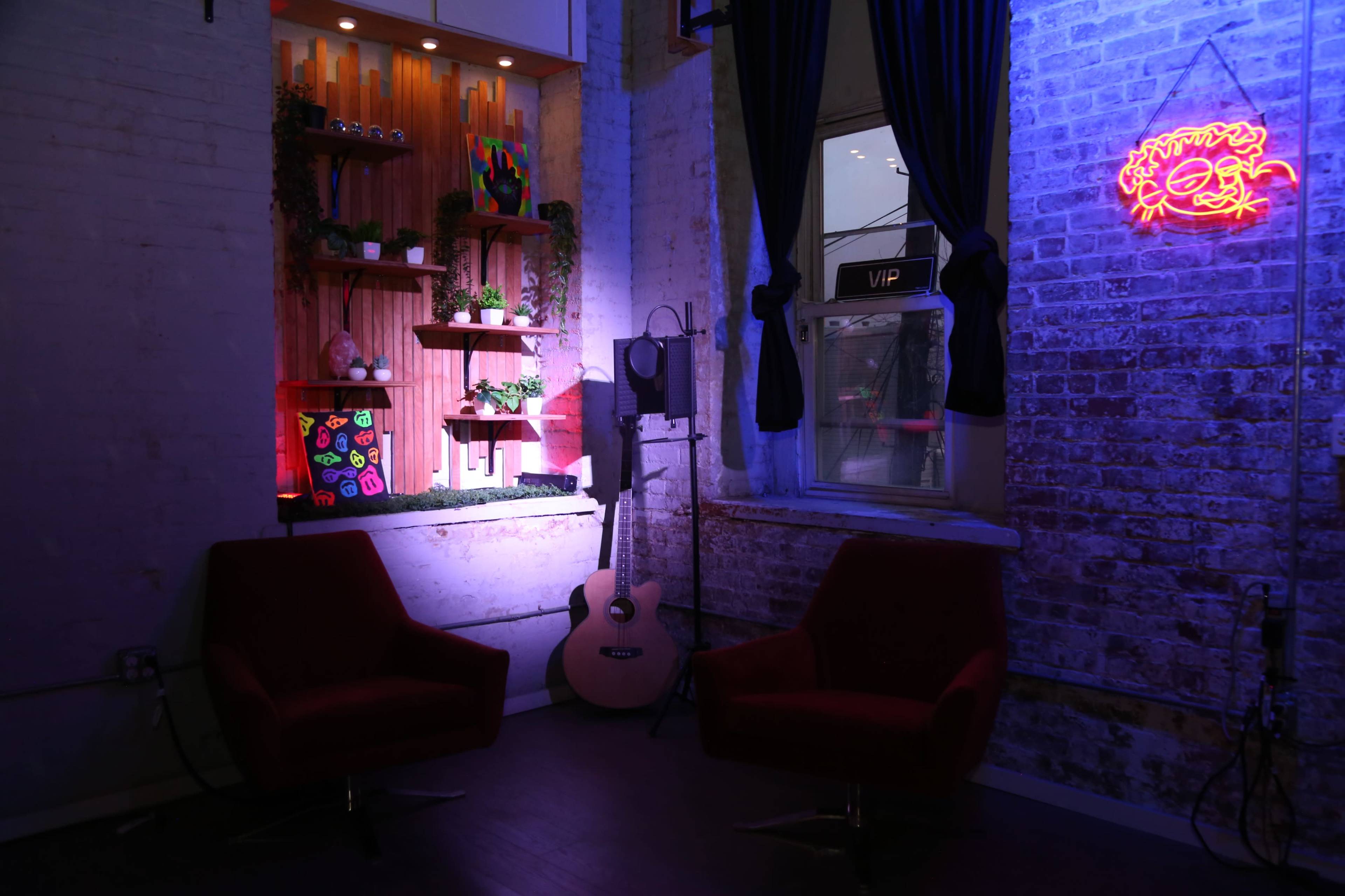 The image shows a cozy corner of a room featuring two red chairs, a wooden shelf with colorful decorations, an acoustic guitar, and a neon sign.
