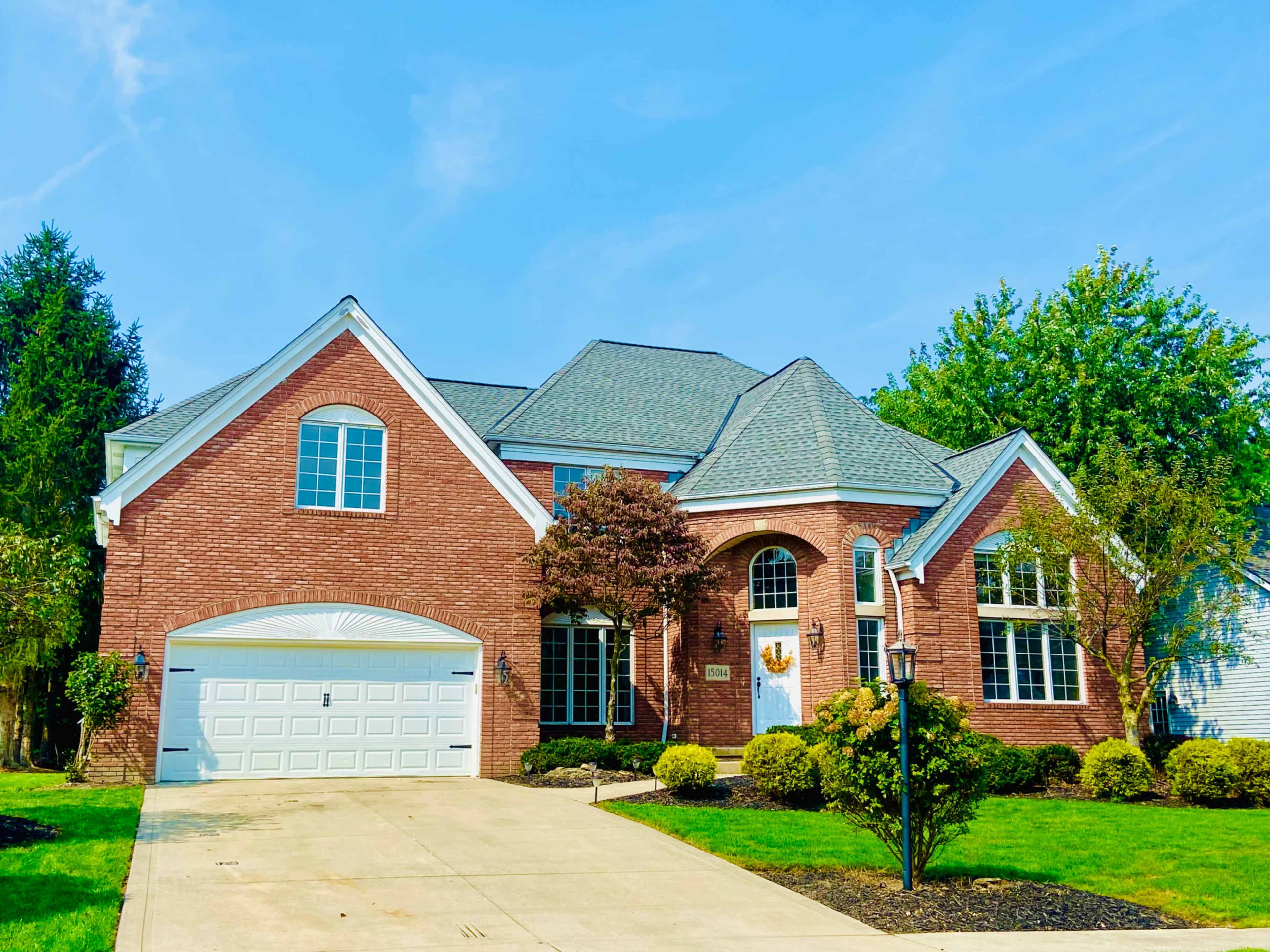 The image shows a large brick house with a manicured lawn, a driveway leading to a garage, and several trees nearby.