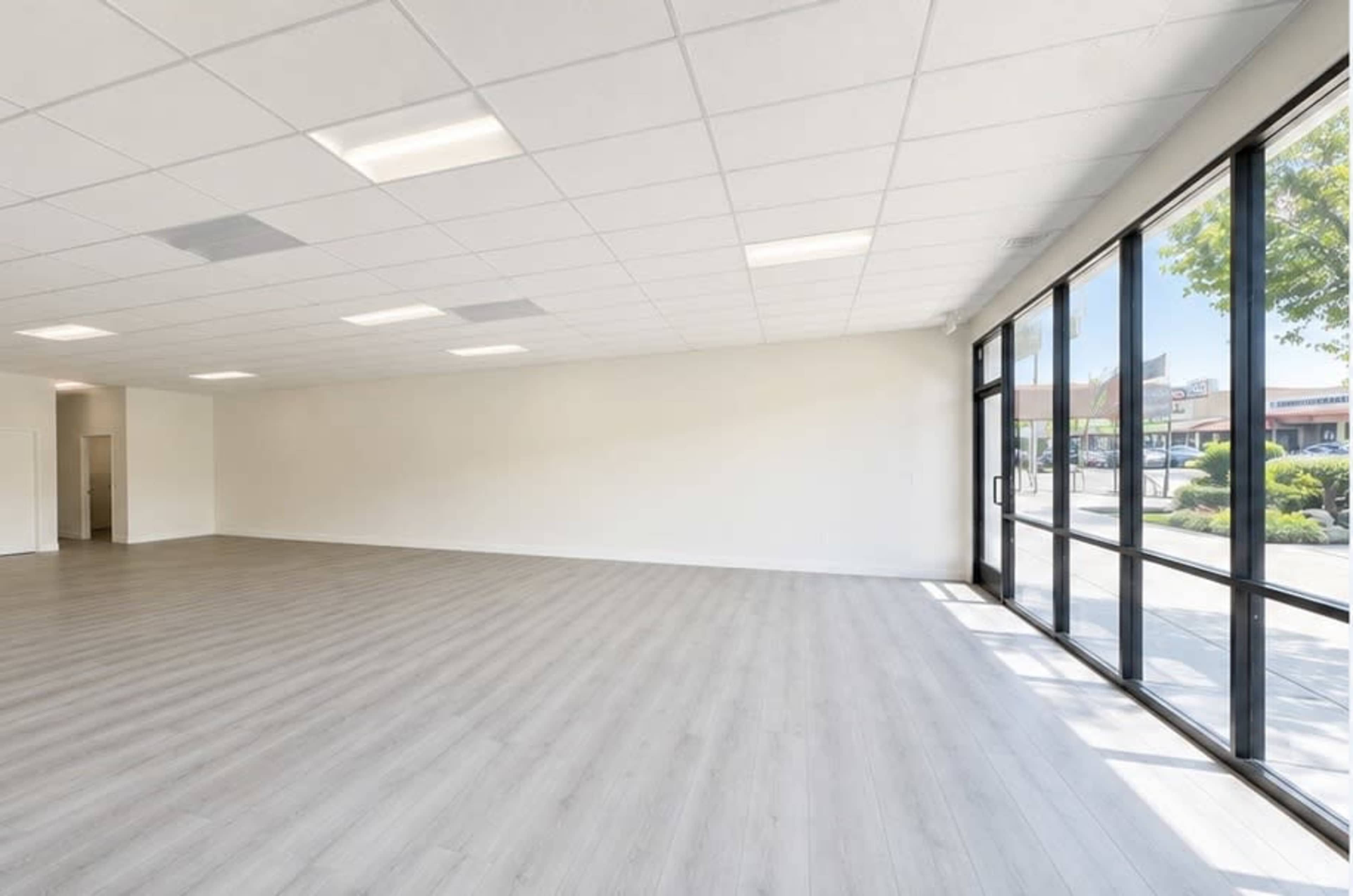 The image shows an empty interior space with a light wood floor, white walls, and large windows letting in natural light.