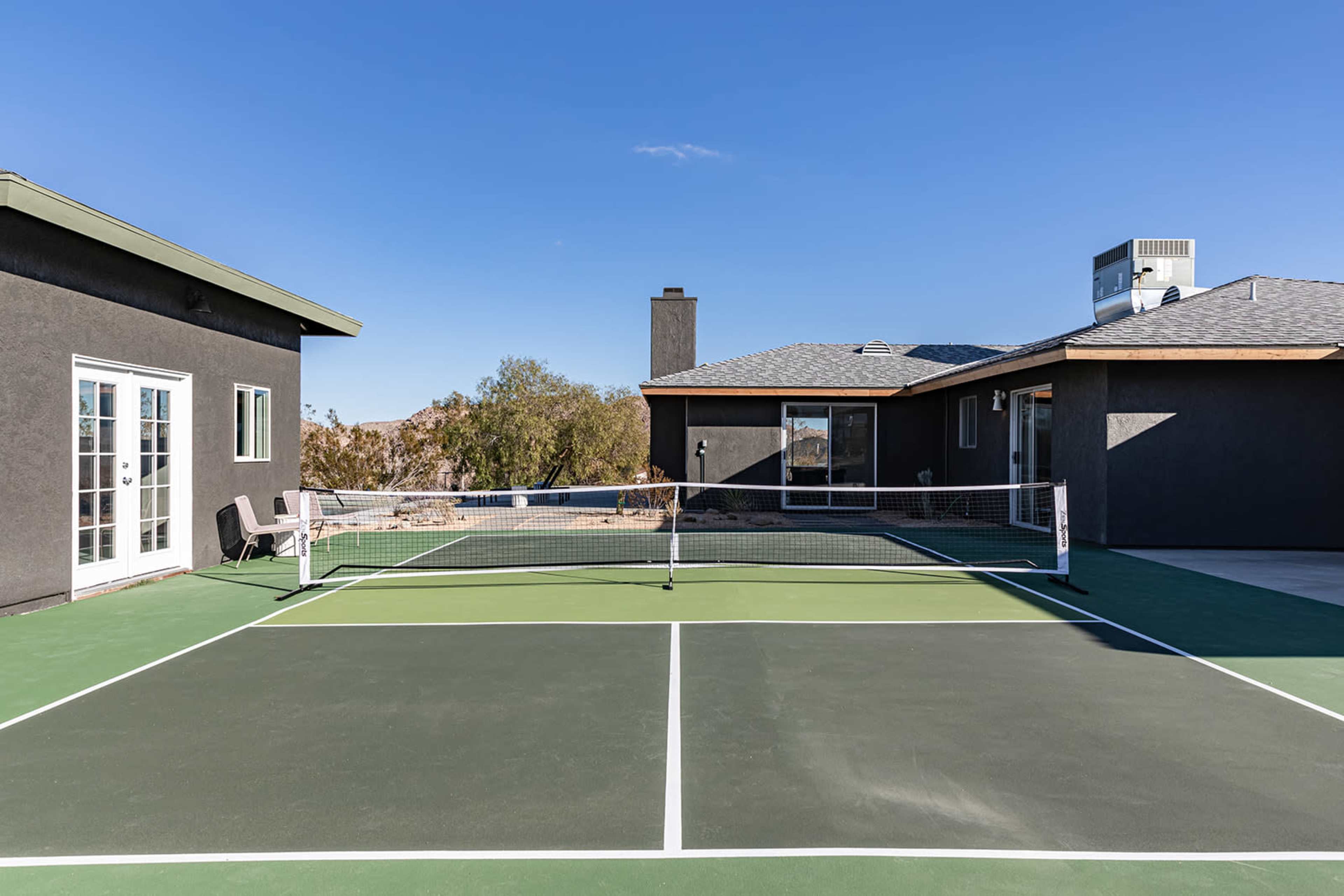 The image shows a green tennis court situated between two dark-colored houses under a clear blue sky.