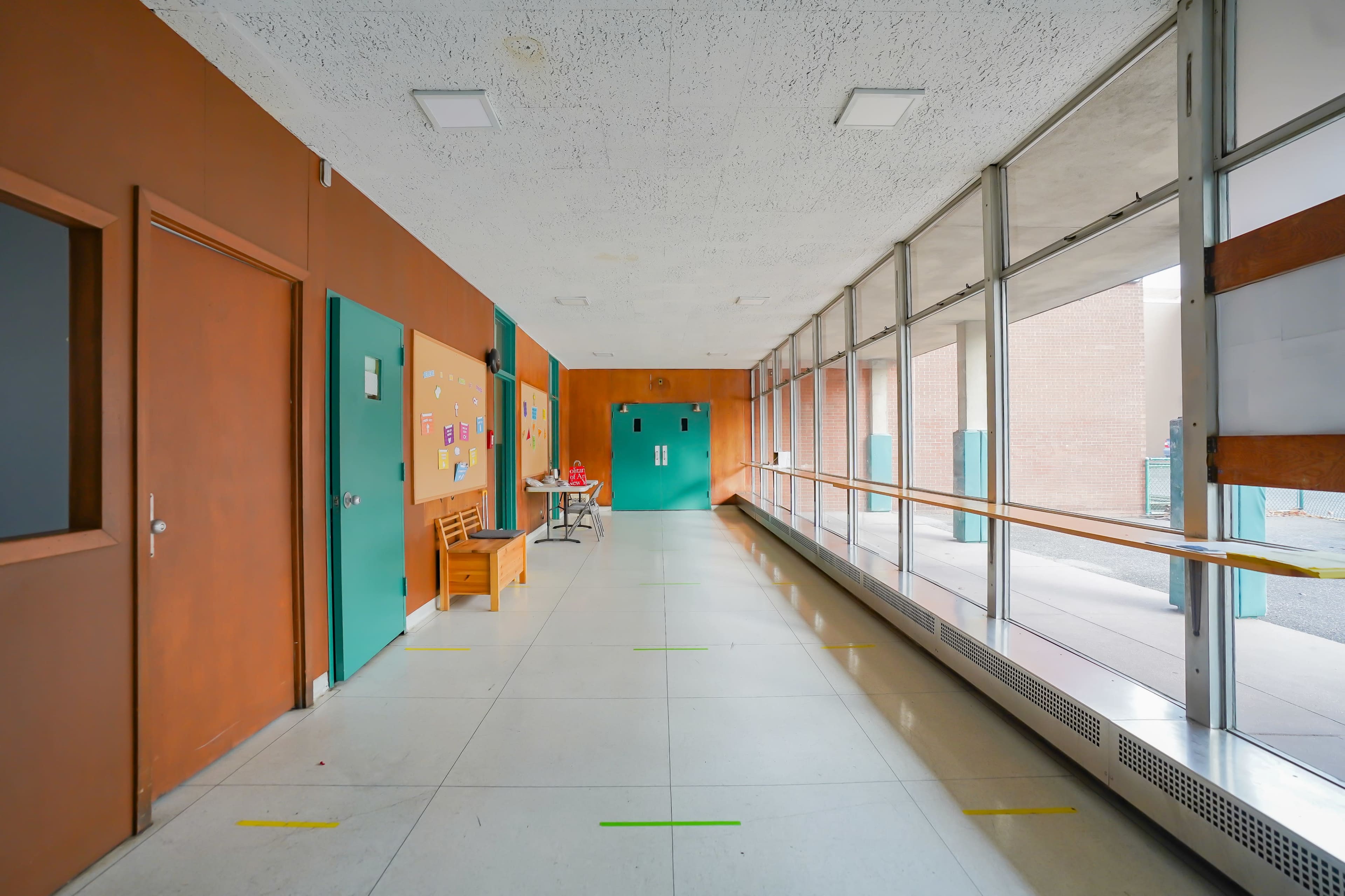 A hallway with walls in orange and green features a bench and large windows on one side, leading to two doors at the end.