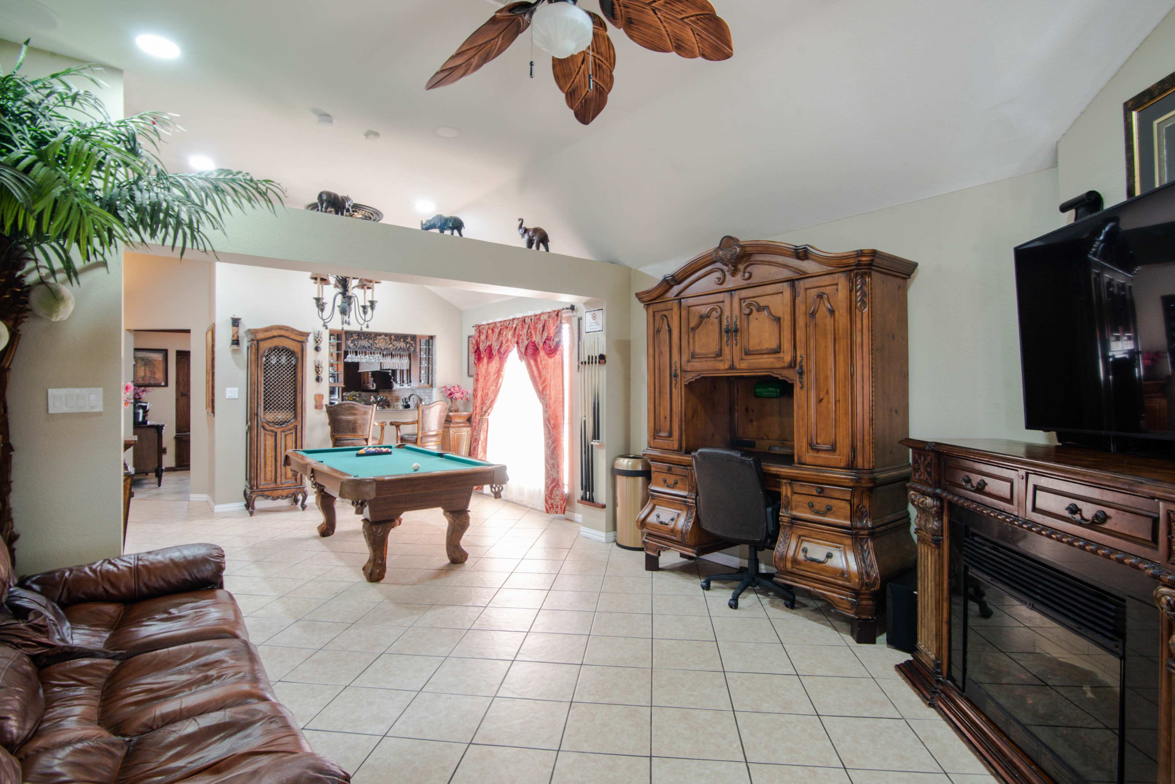 The image depicts a spacious living room featuring a pool table, a wooden desk, and a large entertainment center, with decorative plants and warm lighting.
