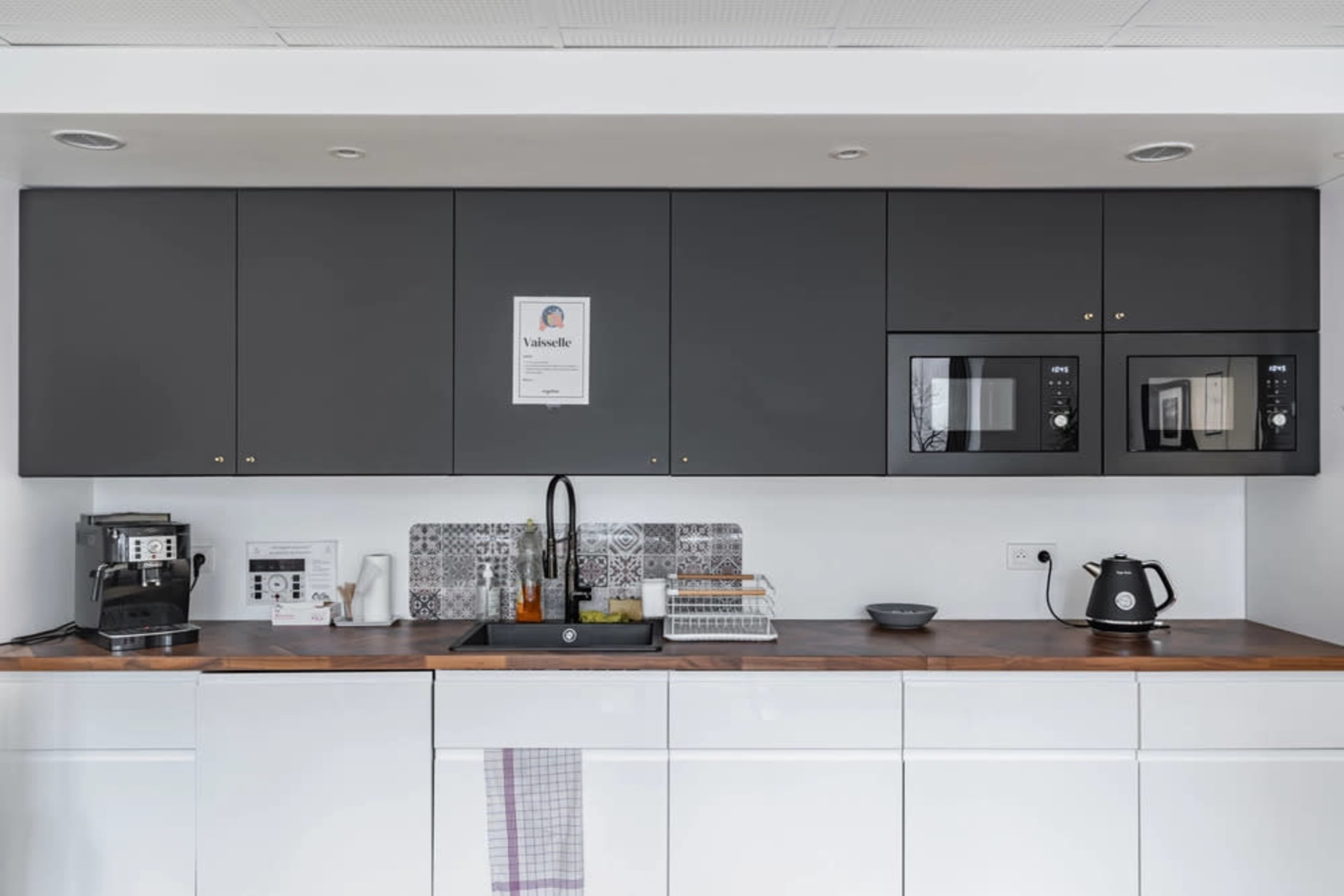 The image shows a modern kitchen with gray cabinetry, two microwave ovens, and a wooden countertop featuring a sink and various kitchen appliances.
