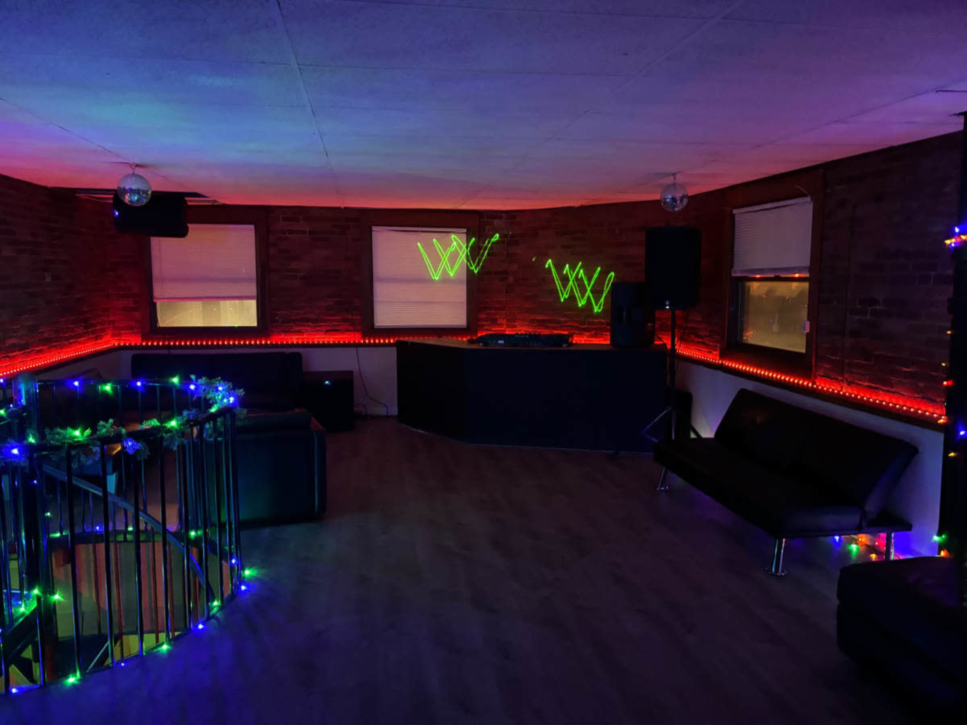 The image shows a dimly lit room with colorful LED lights accentuating a DJ booth and seating areas.