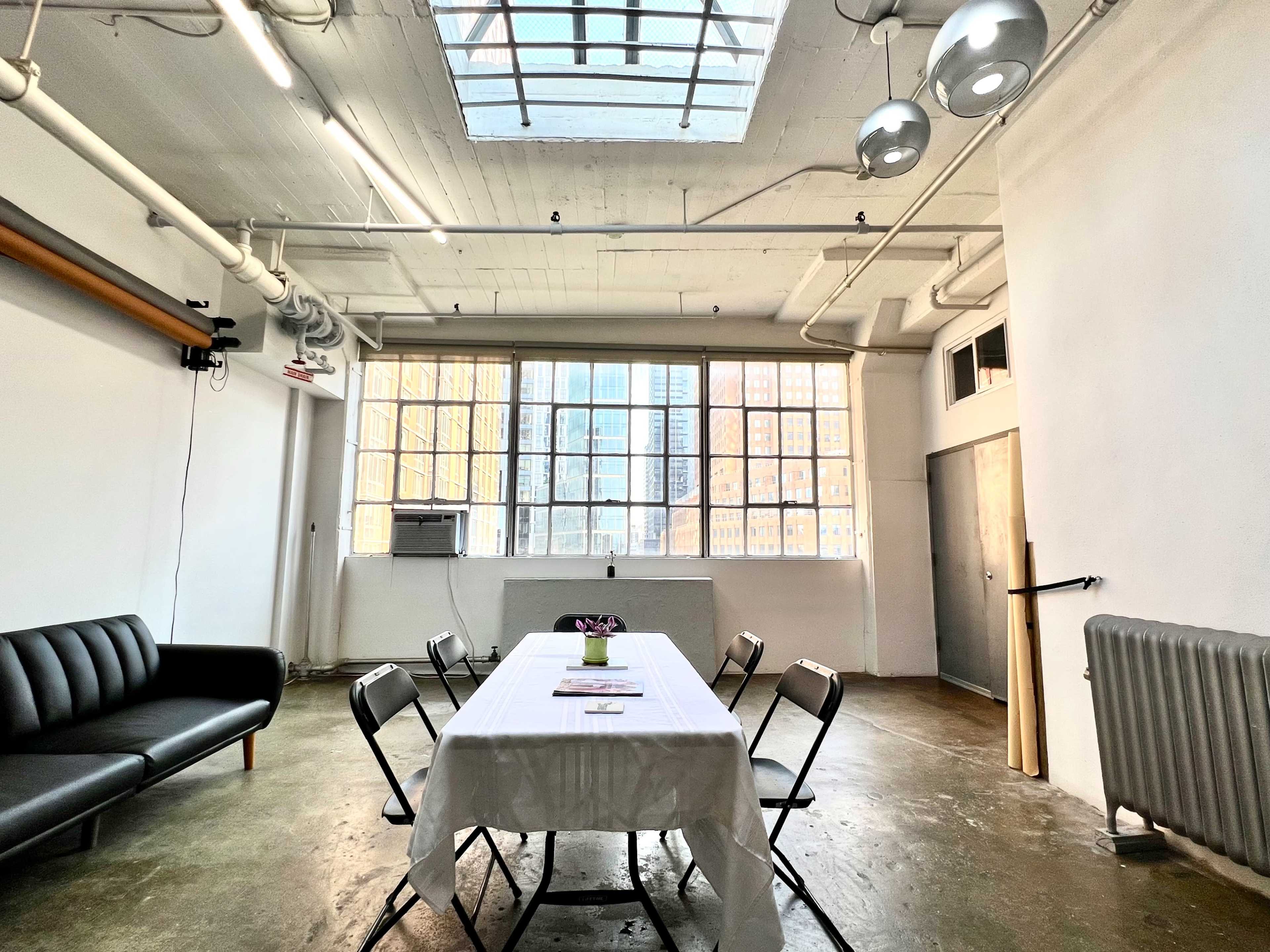 A minimalist meeting room with a long table set for a gathering, a black sofa, large windows allowing natural light, and exposed industrial elements.