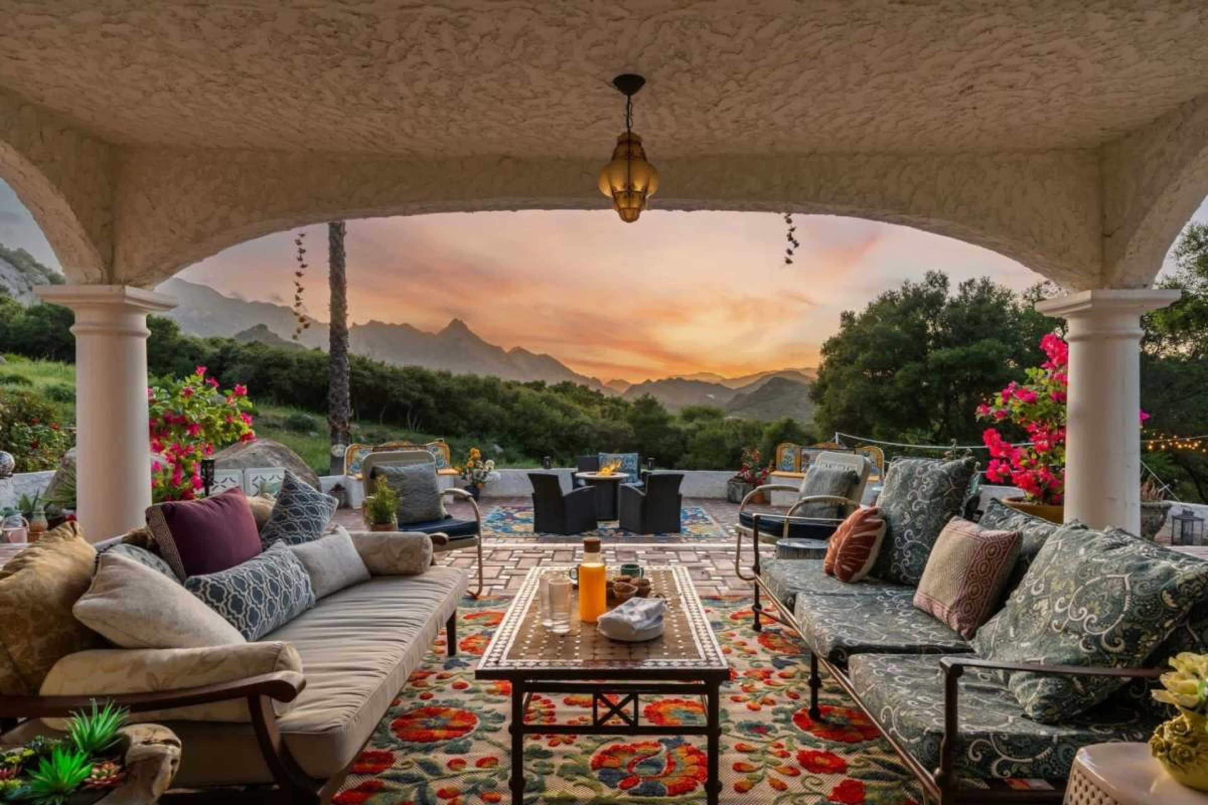 A covered patio is furnished with colorful cushions and a coffee table, overlooking a scenic mountain landscape at sunset.