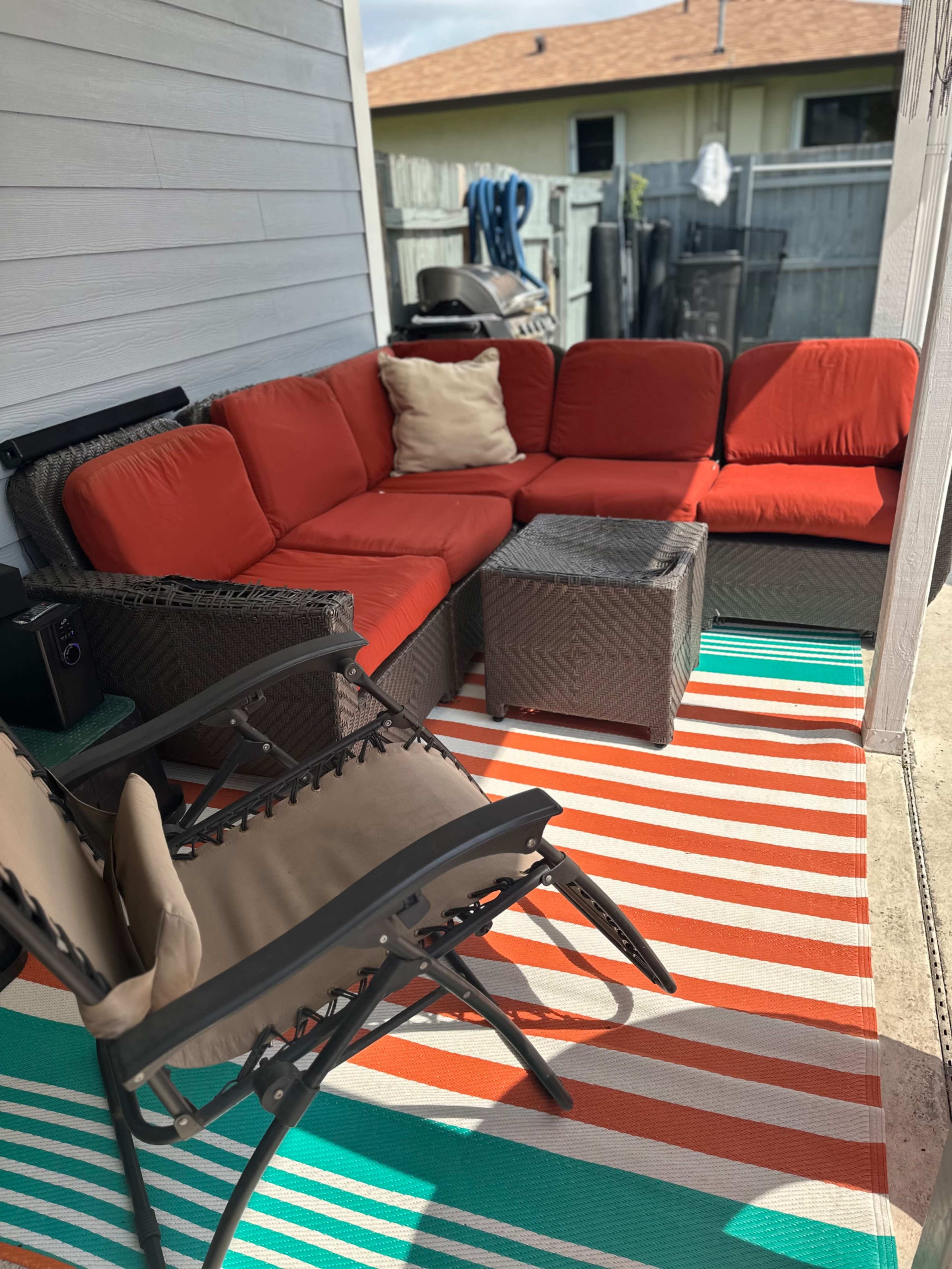 A patio area features a red sectional sofa, a woven coffee table, and a striped rug, with a pair of chairs nearby.