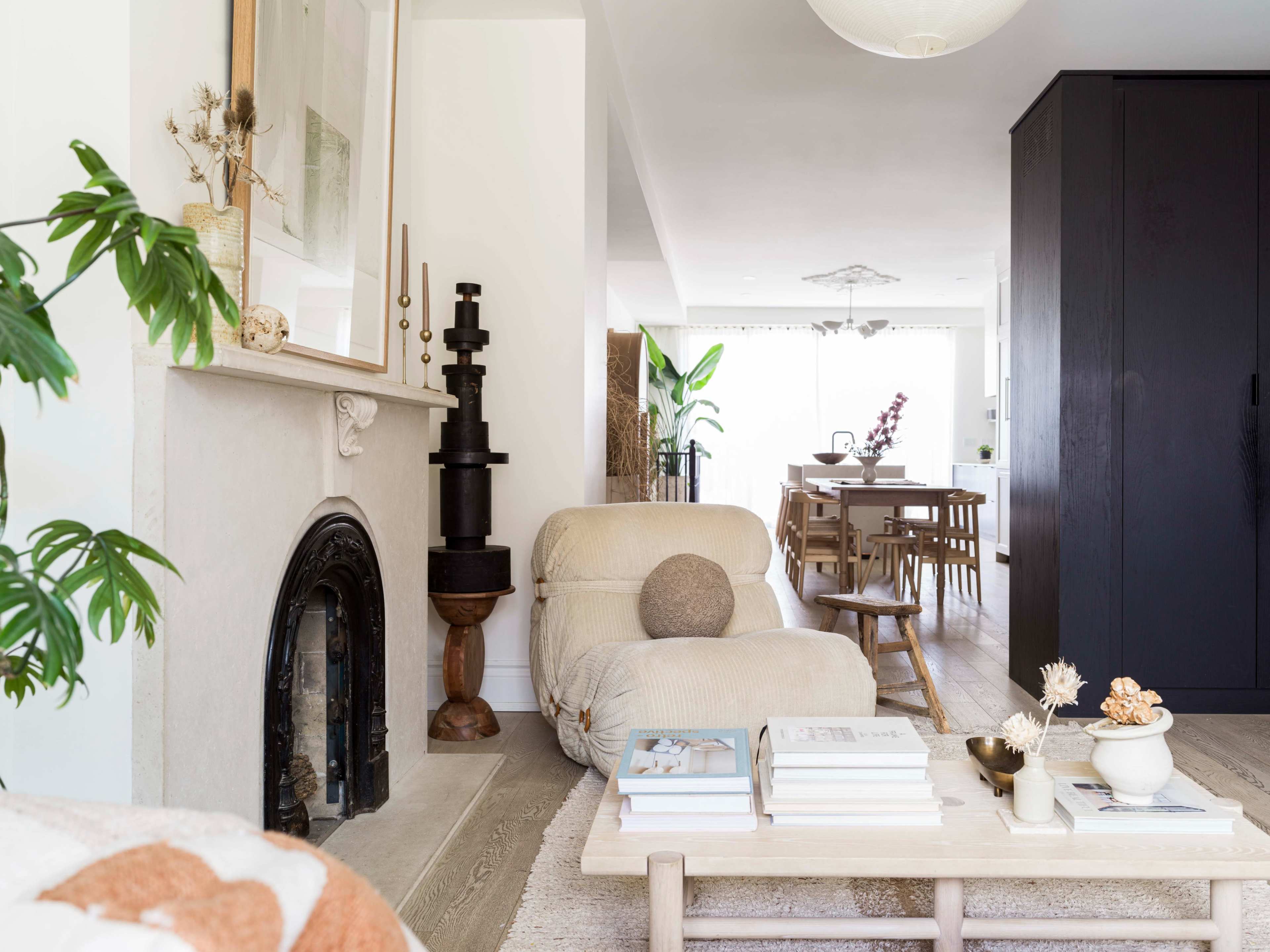 A modern living room with a beige sectional sofa, a coffee table stacked with books, and a dining area in the background featuring wooden furniture.