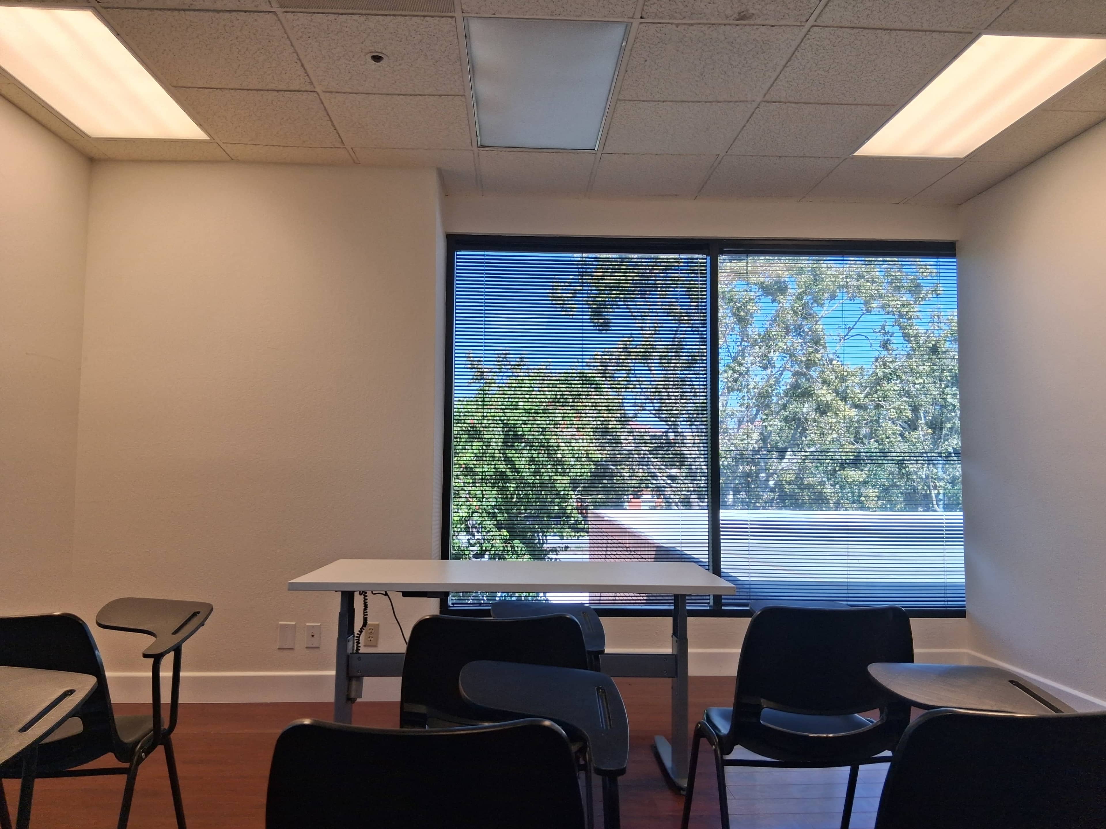 The image shows a light-colored classroom with empty black chairs and a small table, featuring large windows that overlook trees outside.
