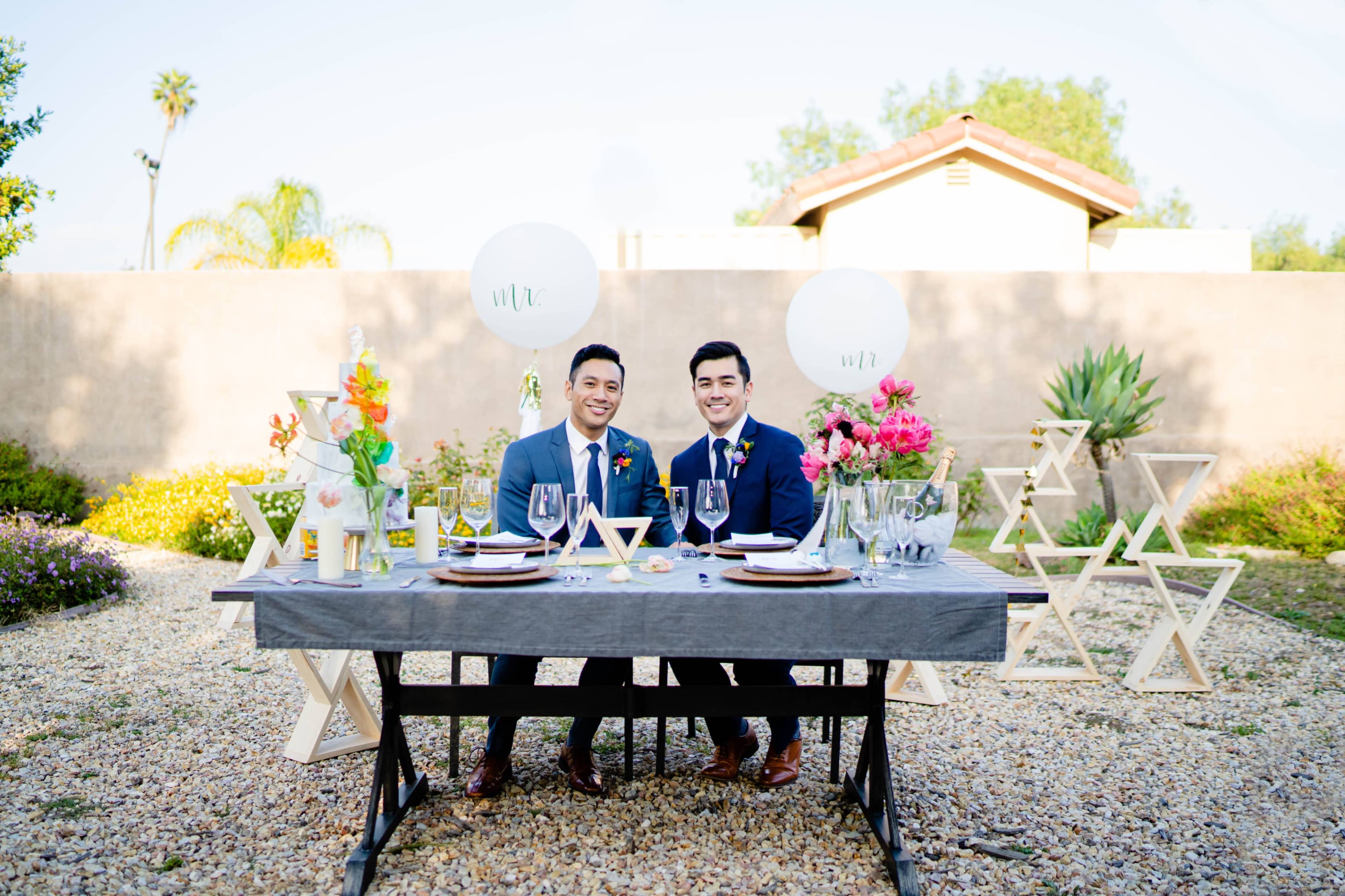 Two men in formal attire sit at a decorated outdoor table set for a celebration, surrounded by flowers and balloons.