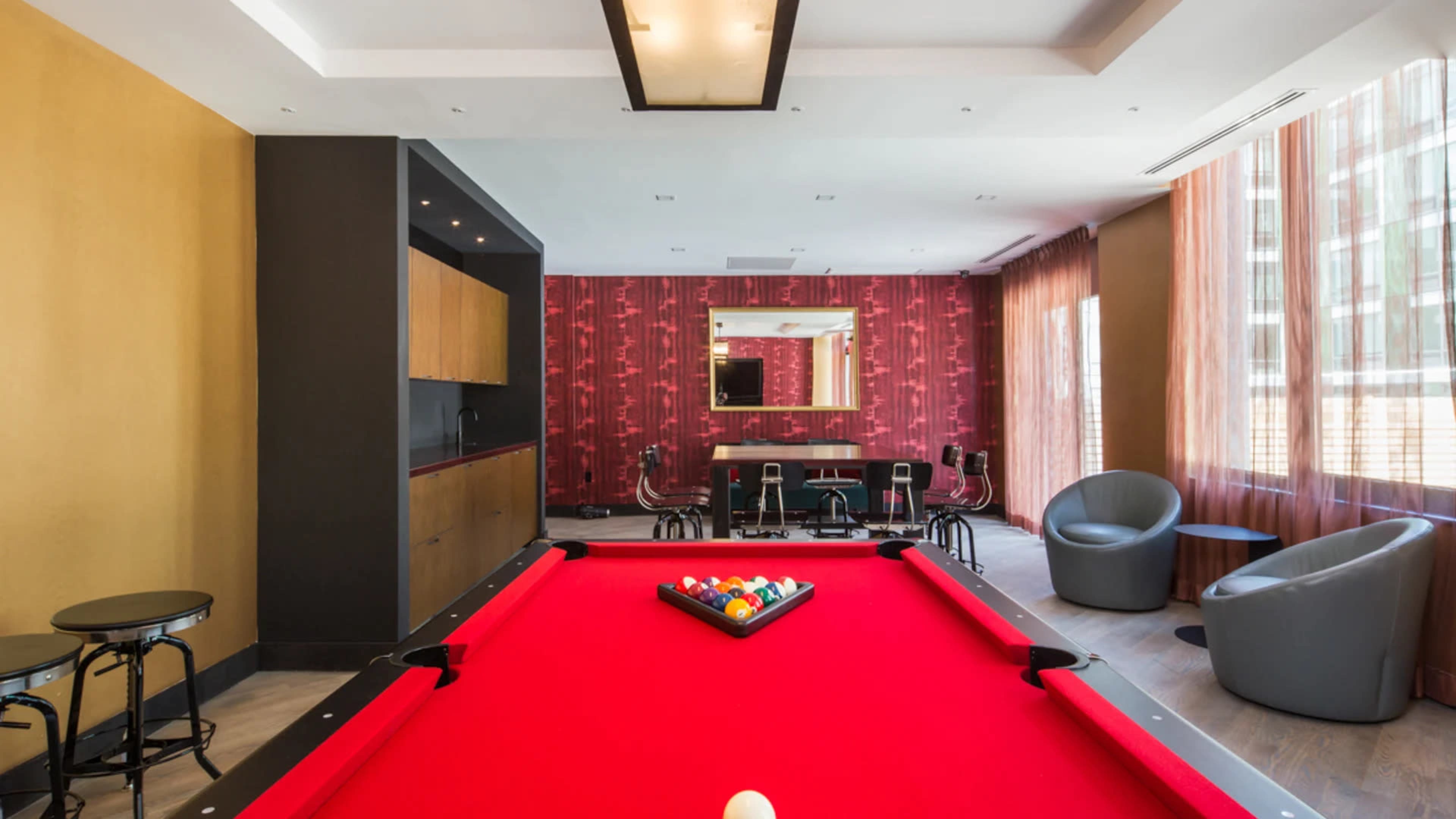 A brightly lit room features a red pool table at the center, surrounded by a bar area and modern seating.