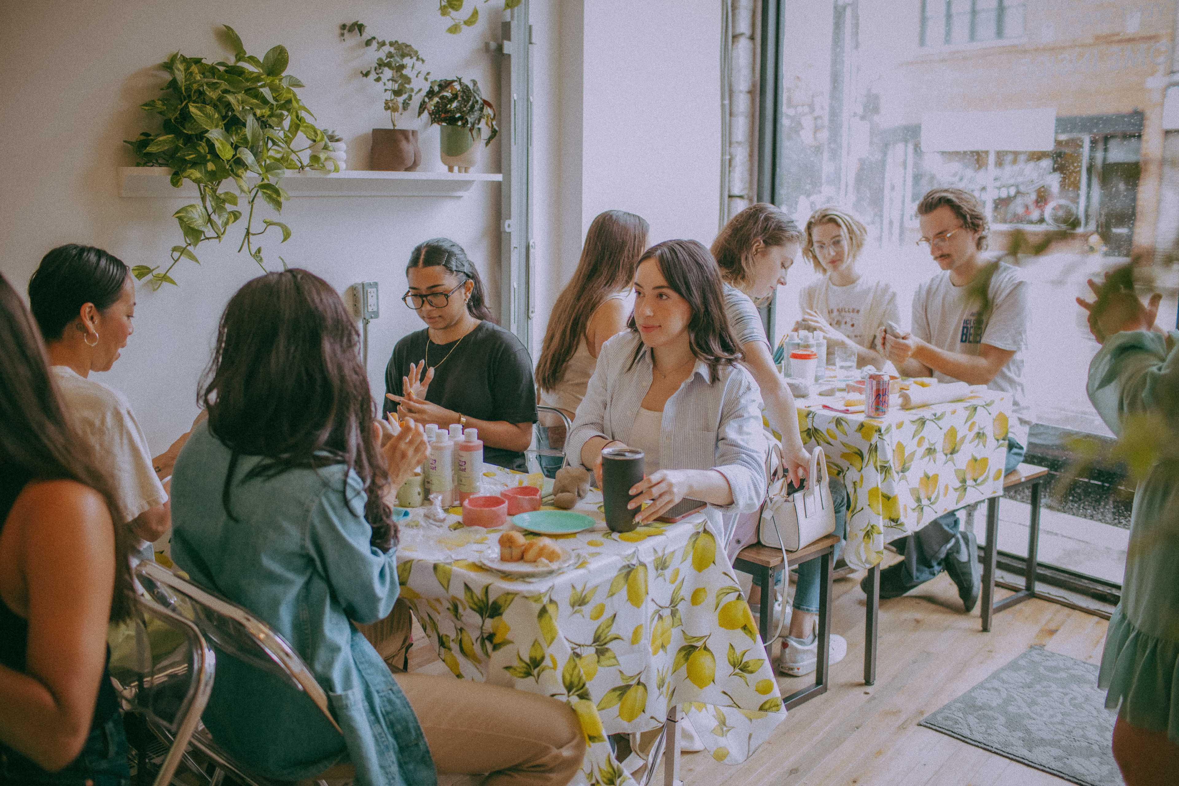 A group of people are gathered around tables adorned with lemon-patterned tablecloths, engaging in a craft activity inside a bright café with large windows.