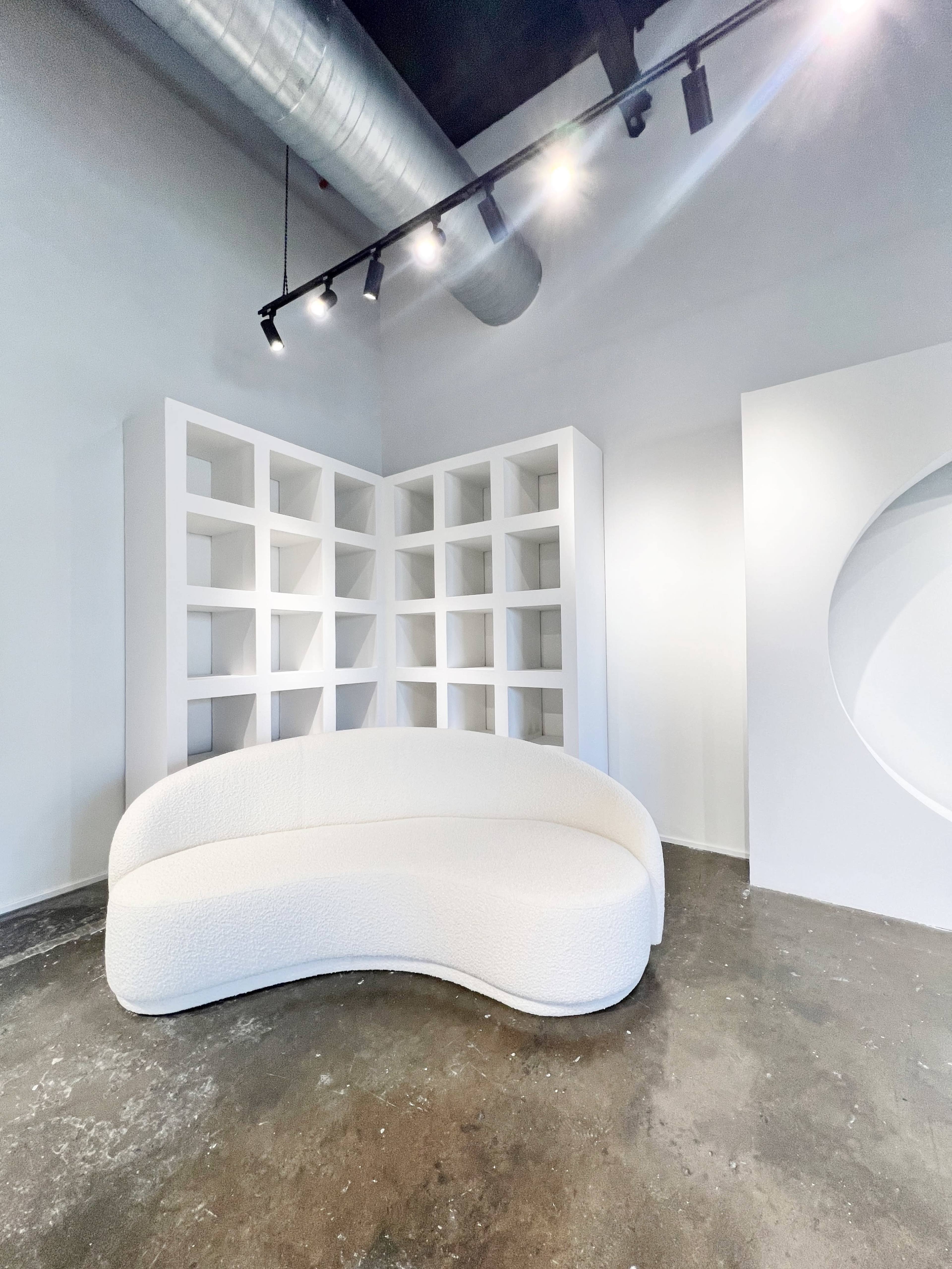 The image features a modern white curved sofa positioned in front of a bookshelf with open cubed spaces, set against a minimalist interior.