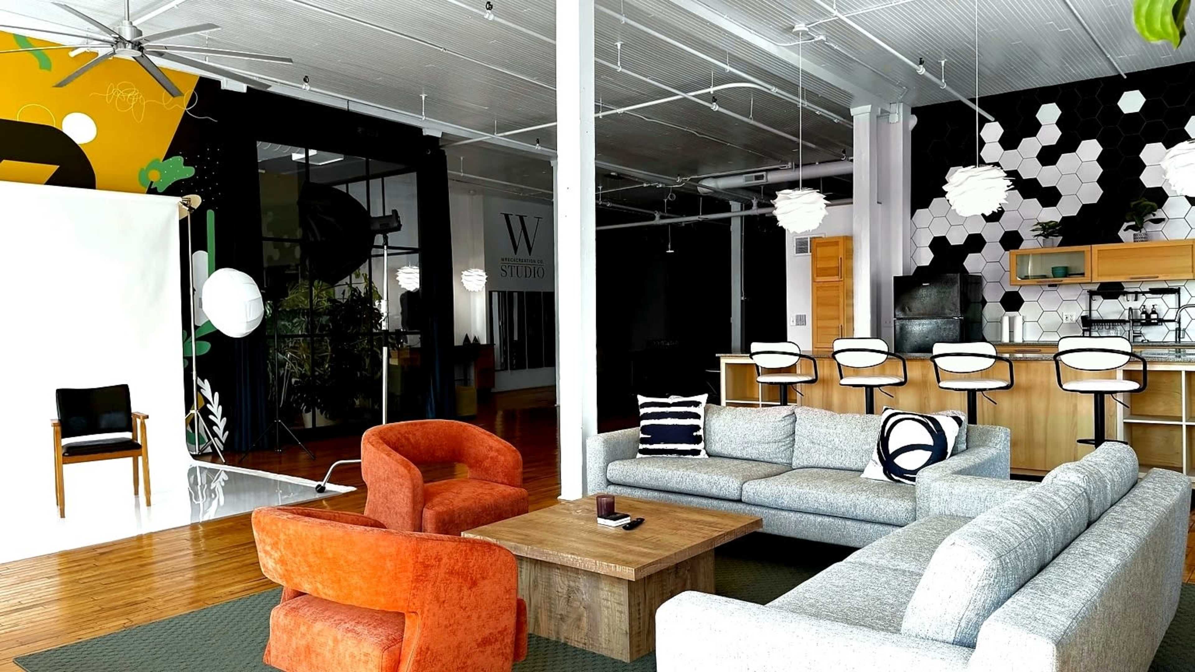 The image shows a modern studio space featuring a mix of seating options, a large wooden table, and a kitchen area with bar stools, all set against a backdrop of contrasting black and vibrant wall designs.