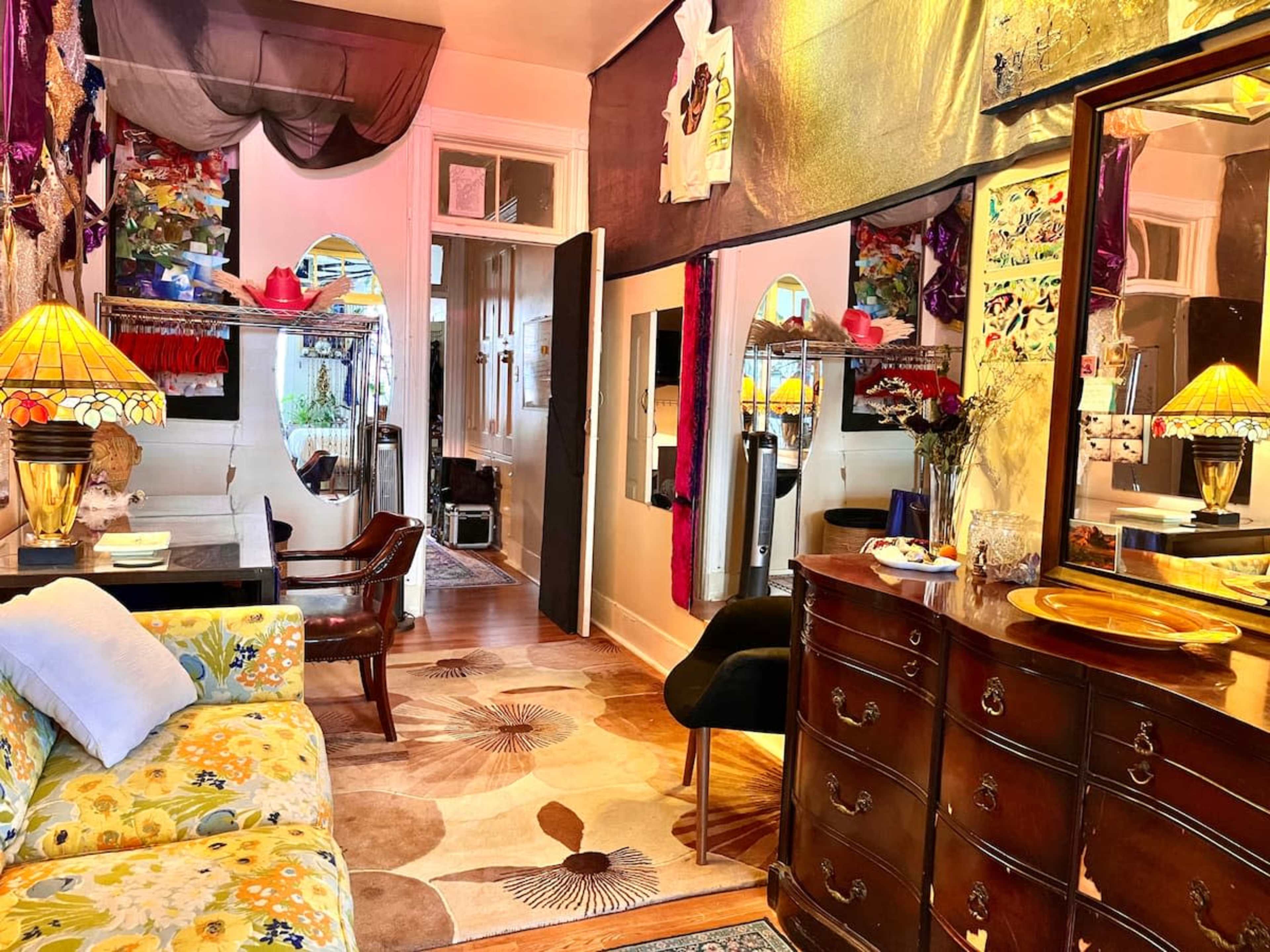 The image shows a vibrant, eclectic room featuring floral-patterned furniture, a decorative rug, and various artistic elements on the walls.