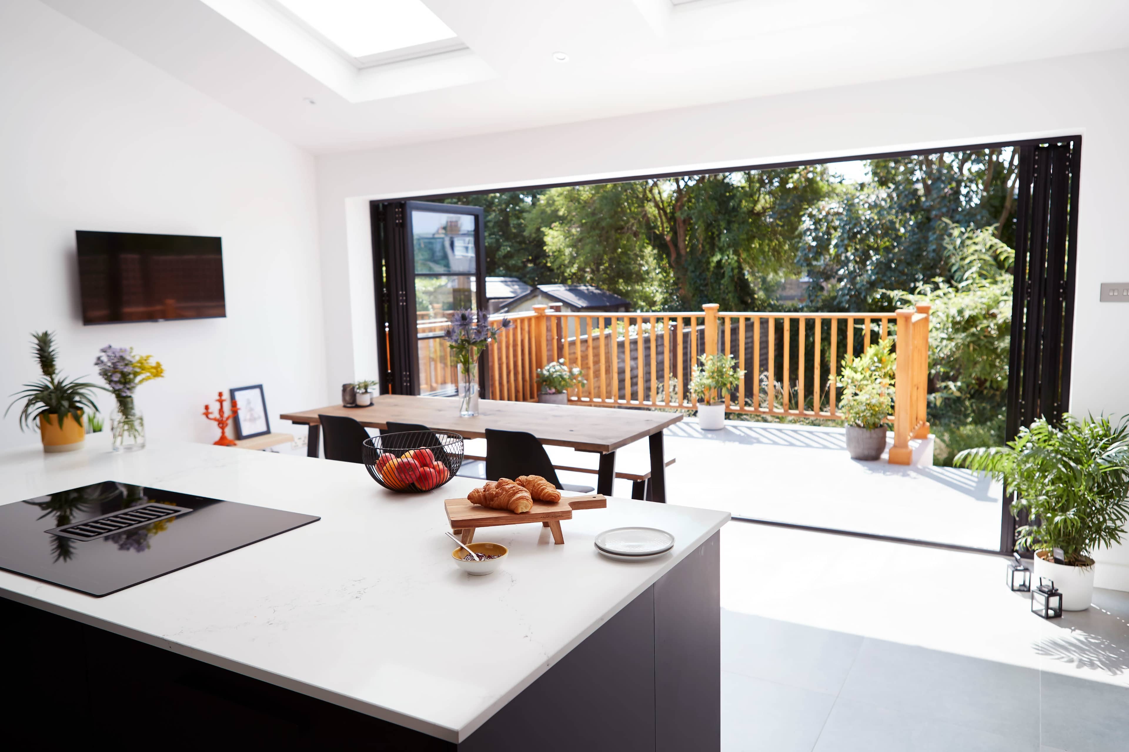 A modern kitchen with an island counter, leading to a dining area that opens onto a wooden deck surrounded by greenery.