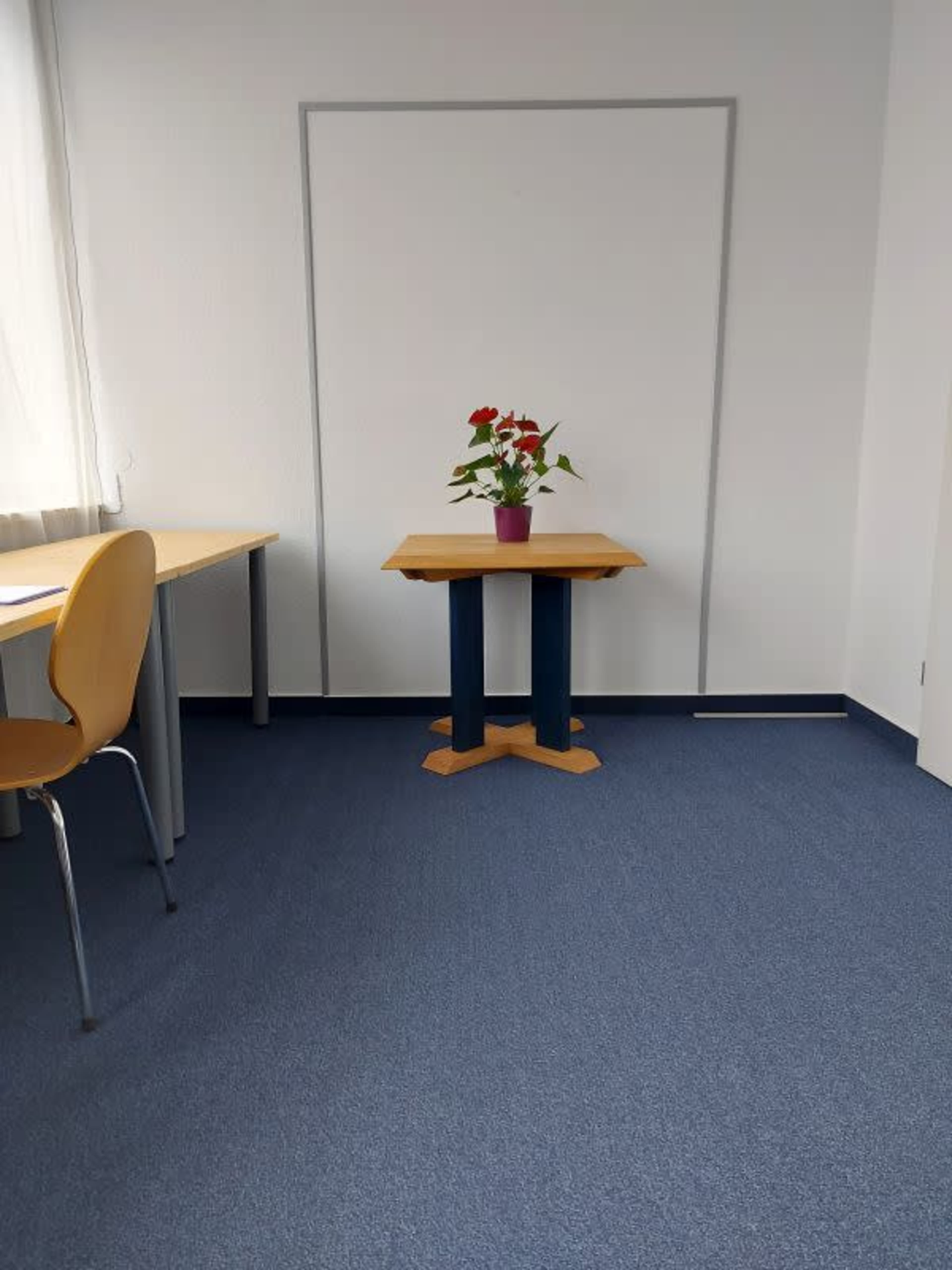 A small room features a table with a potted plant and a chair, with a plain wall and another empty table visible in the background.