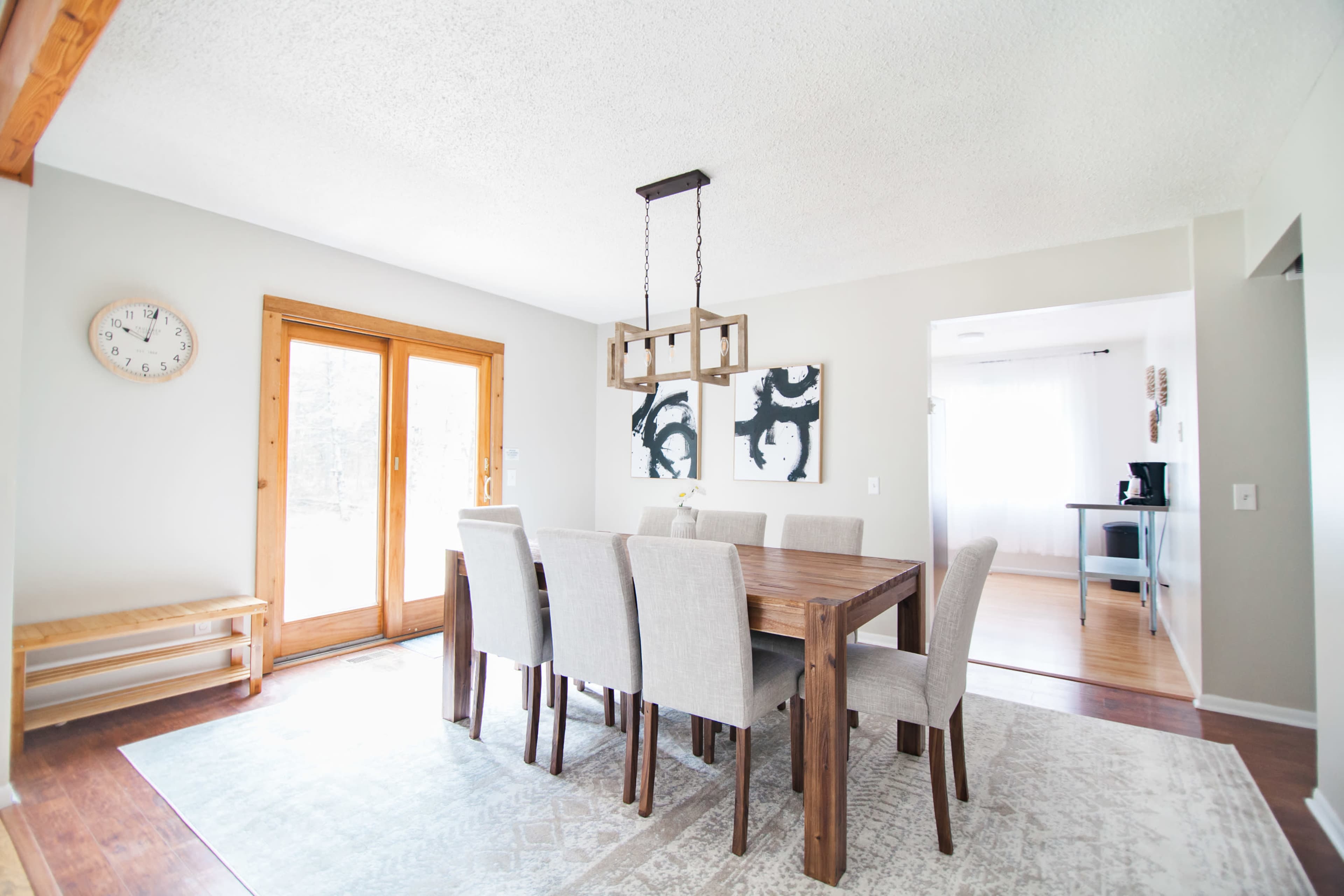 A dining room features a wooden table surrounded by upholstered chairs, large windows, and a light fixture overhead.