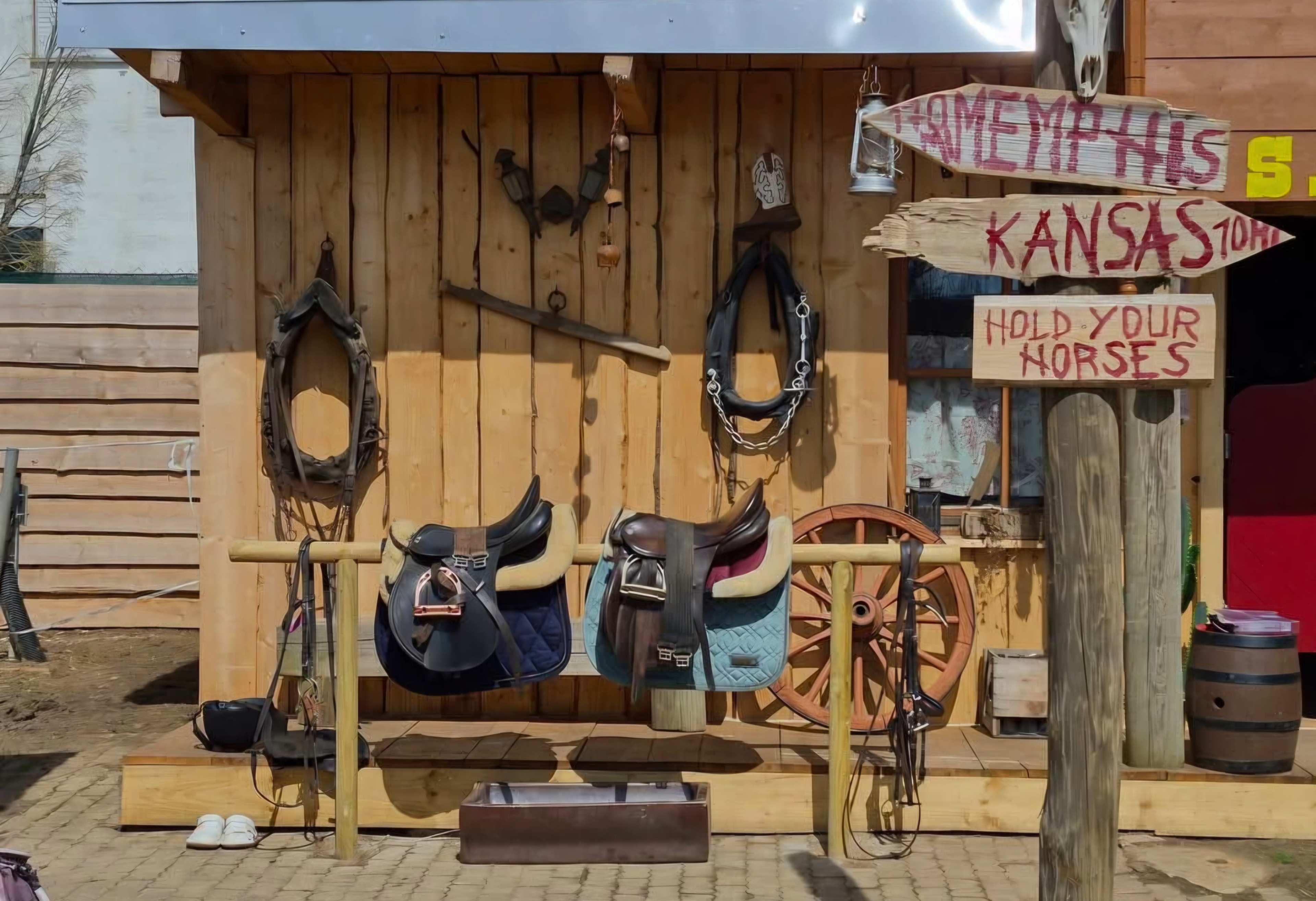 The image shows a wooden wall display with various horseback riding saddles, reins, and a large wooden wheel, along with a sign reading "Welcome to Kansas, Hold Your Horses."