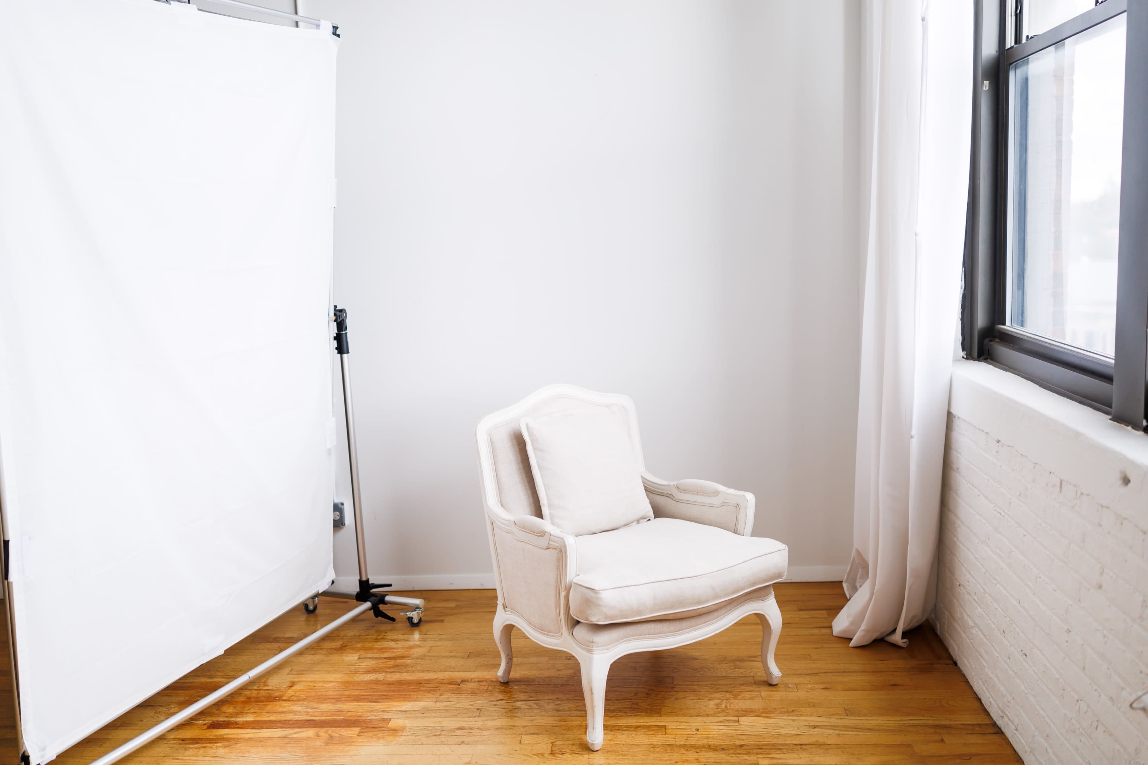 A white upholstered chair is positioned beside a large window in a bright, minimalistic room.