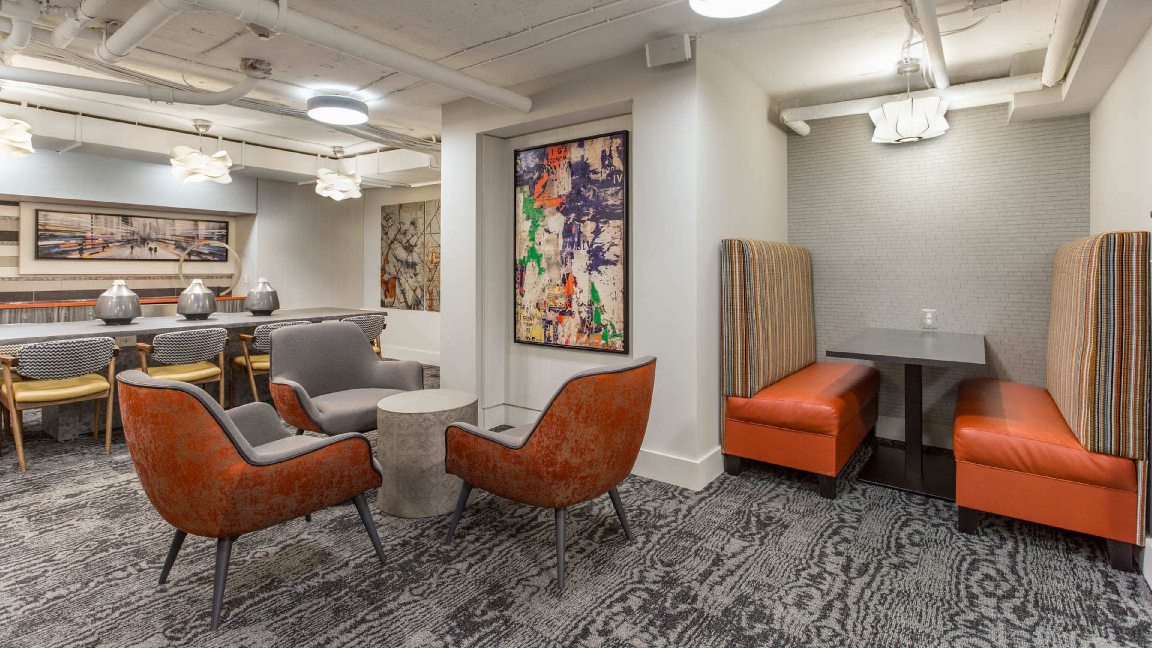 A modern lounge area with a mix of seating options, including two orange armchairs, a gray sofa booth, and a communal dining table.