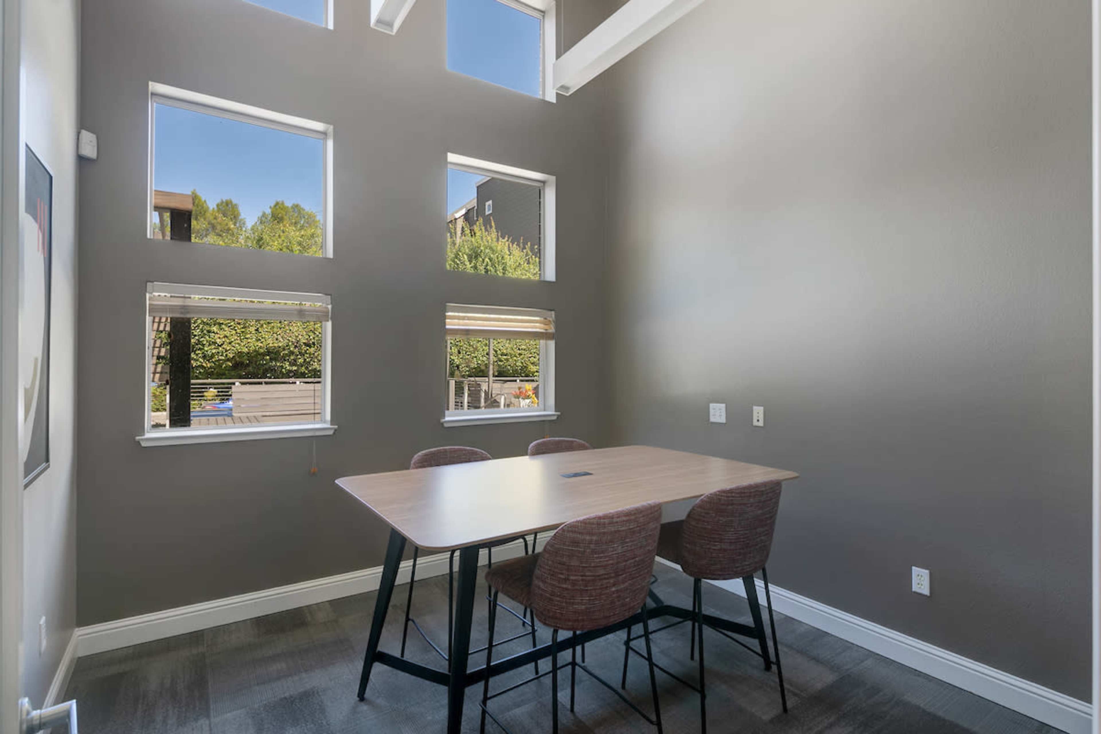 A dining area features a wooden table surrounded by chairs, illuminated by natural light streaming through multiple windows.