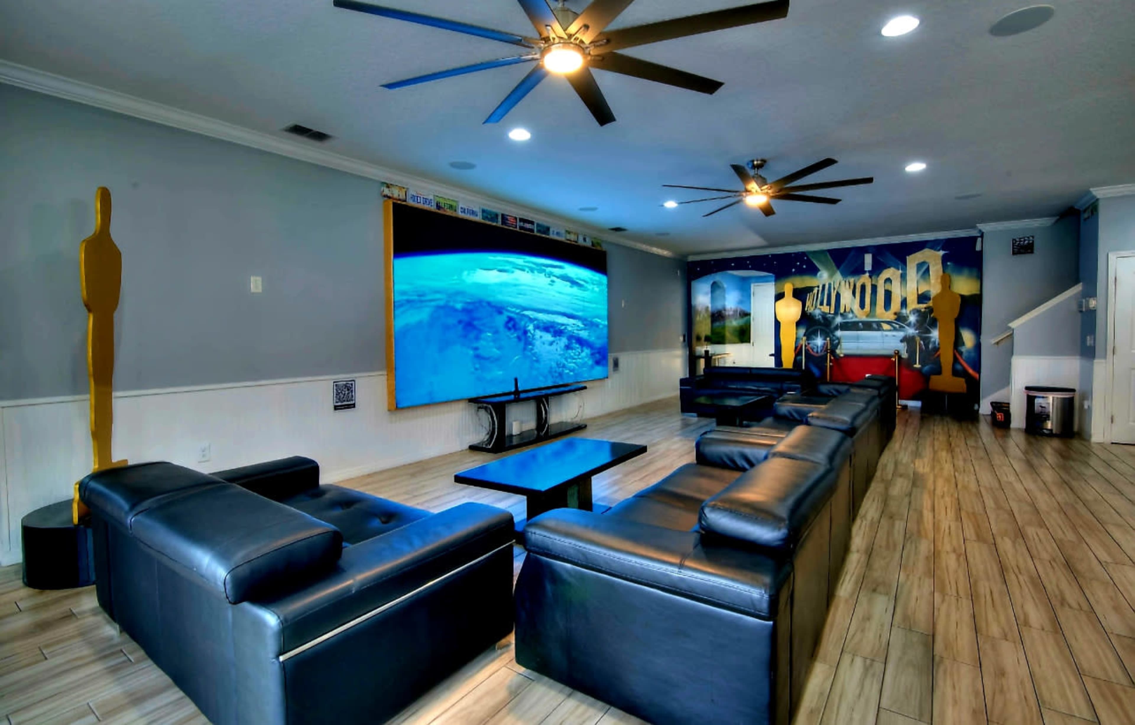 A spacious living room features dark leather seating arranged around a central coffee table, with a large screen displaying a blue image and a mural on the adjacent wall.