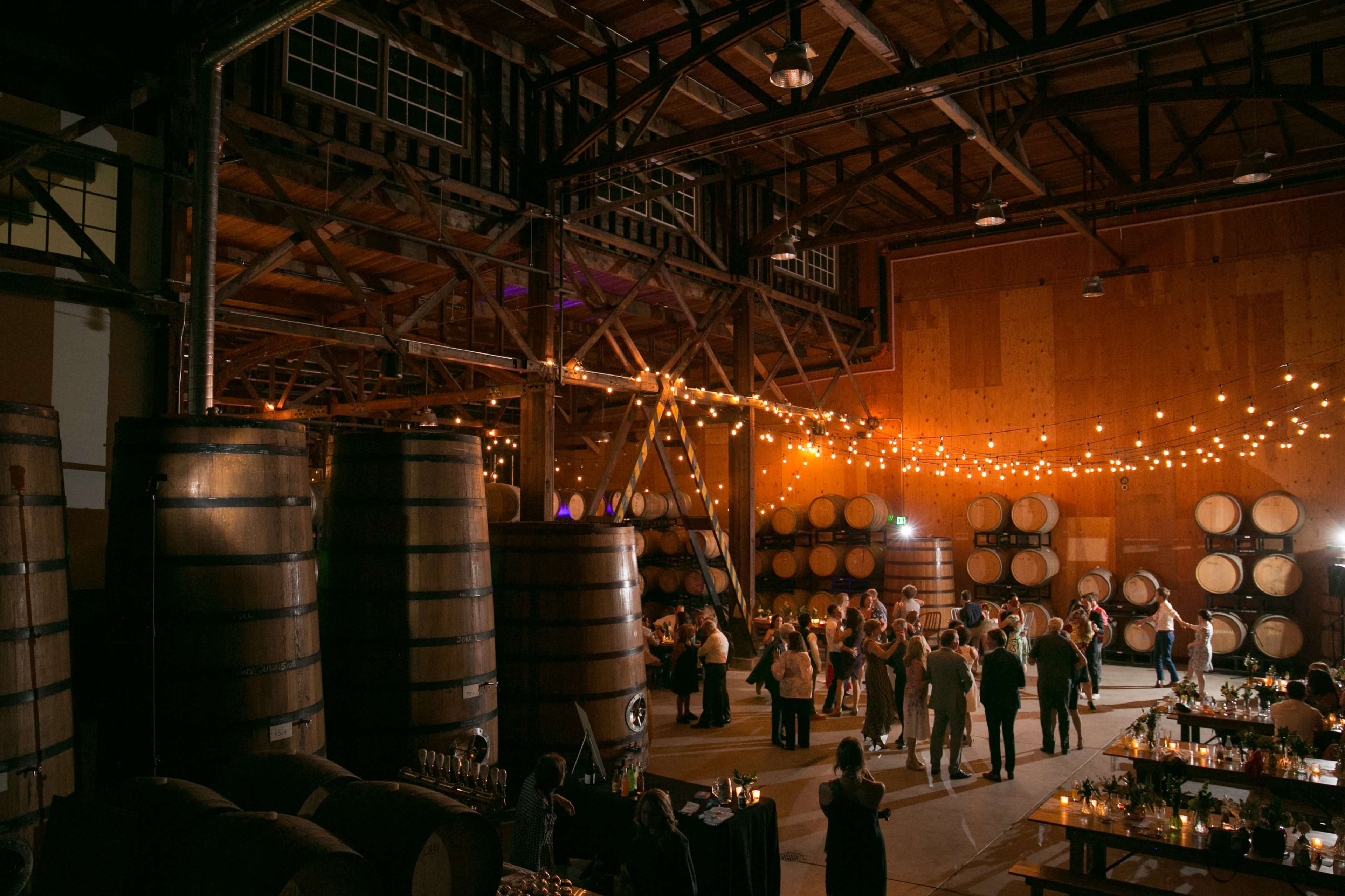 A large gathering of people is taking place in a rustic warehouse setting filled with wine barrels and string lights.