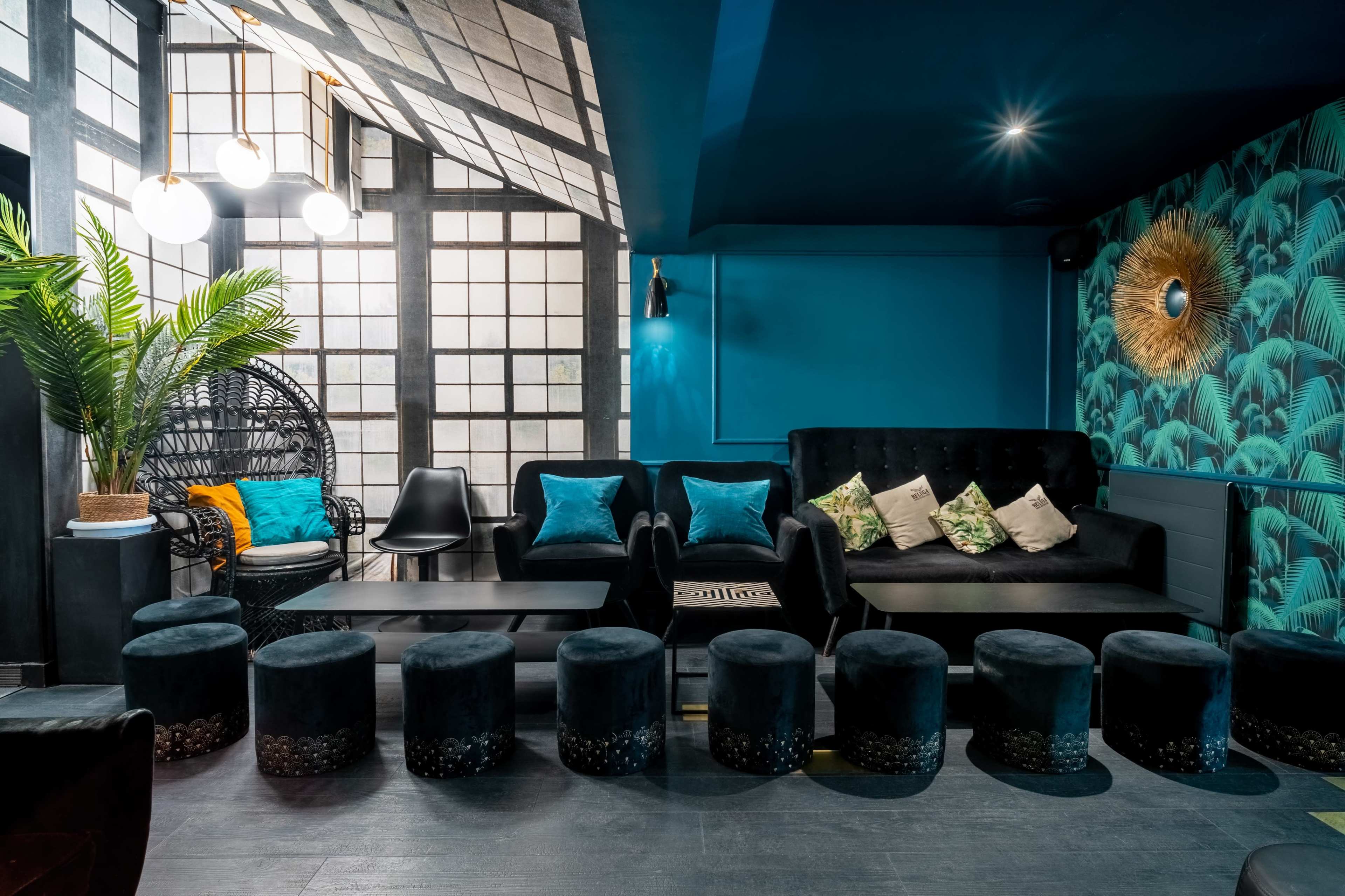 A stylish lounge area with a black leather sofa, colorful pillows, and round stools arranged around a central table, complemented by large windows and tropical-patterned wallpaper.