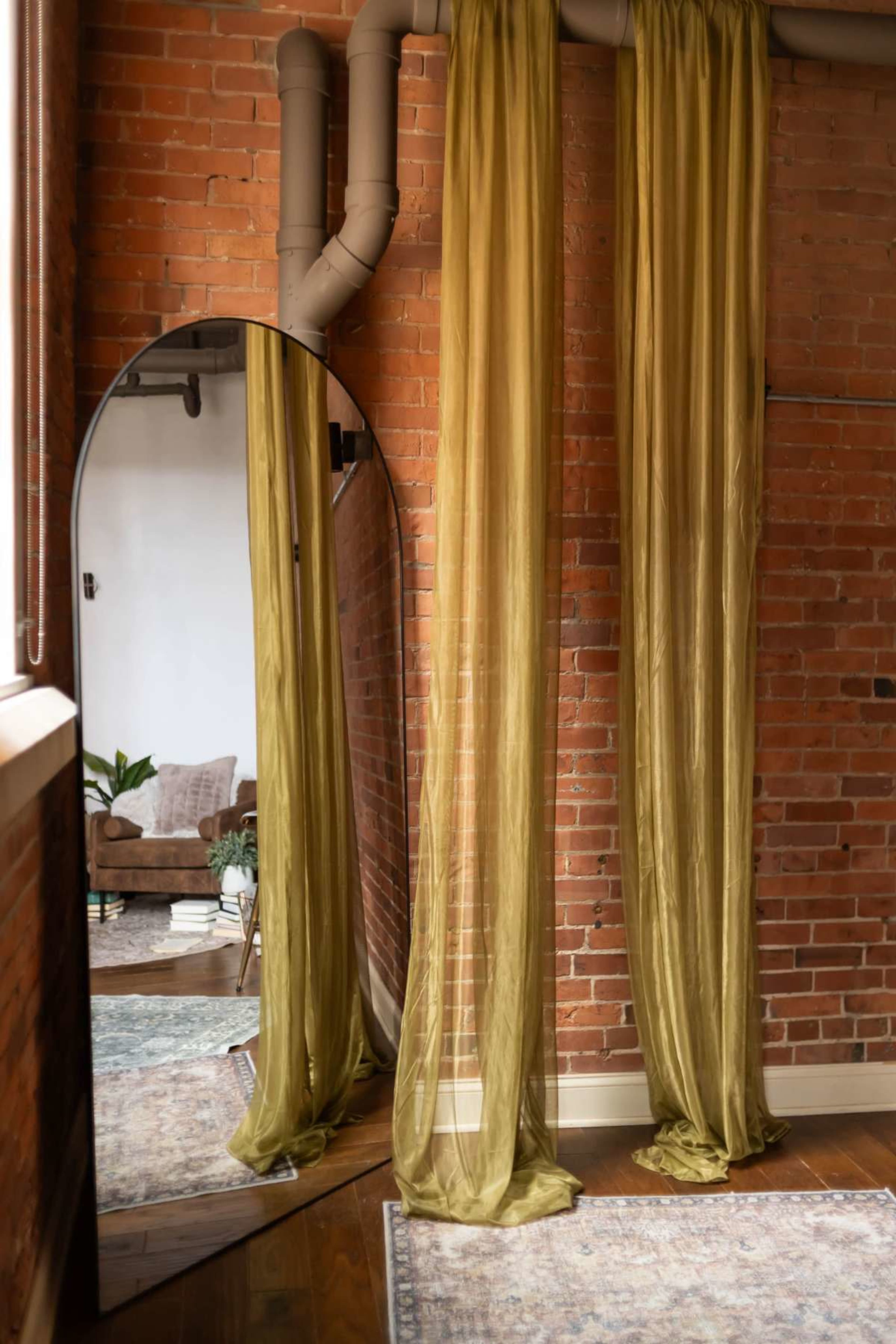 The image features sheer olive green drapes hanging beside a round mirror, with exposed brick walls and a glimpse of a cozy living room in the background.