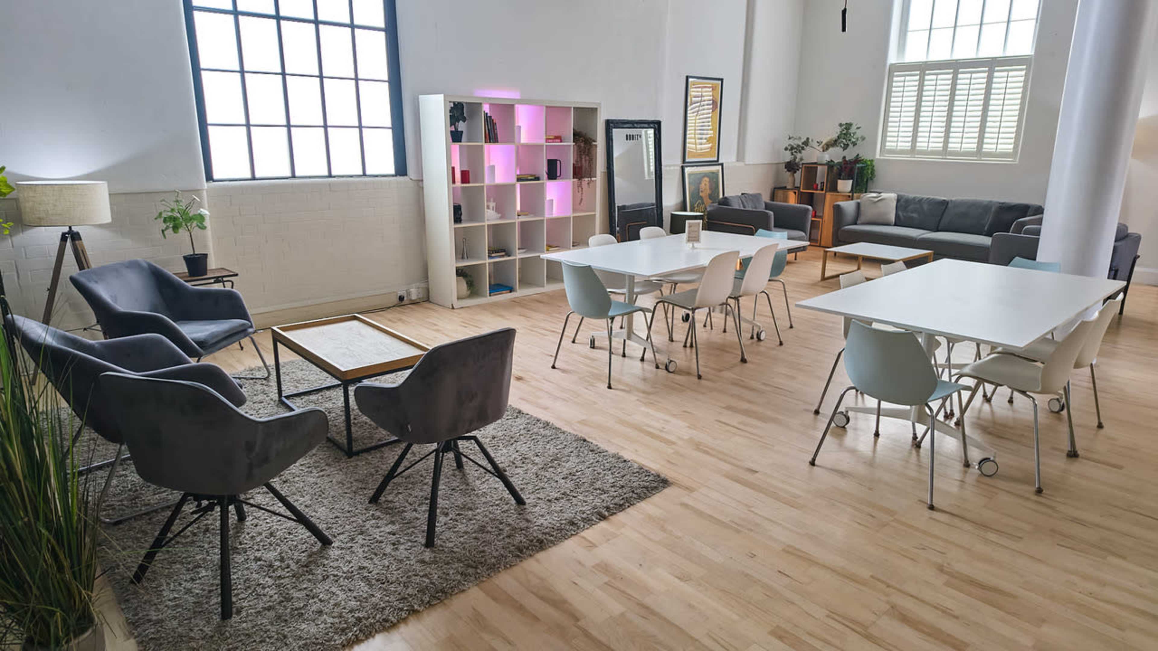 The image shows a modern, airy co-working space with a combination of seating areas, tables, and a bookshelf against the wall.