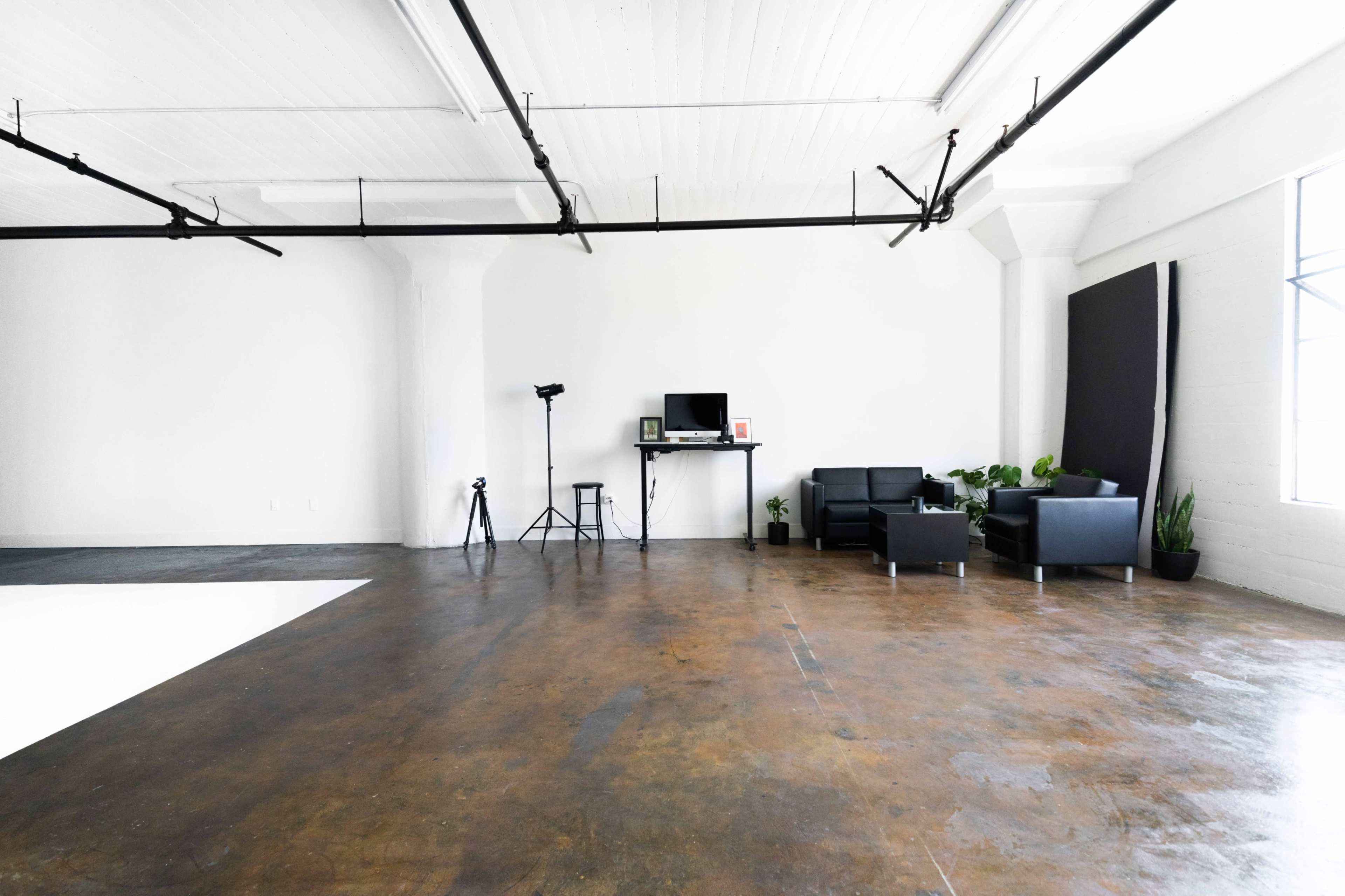 A spacious, minimalist studio with polished concrete flooring, two black chairs, a desk with a computer, and a camera setup.