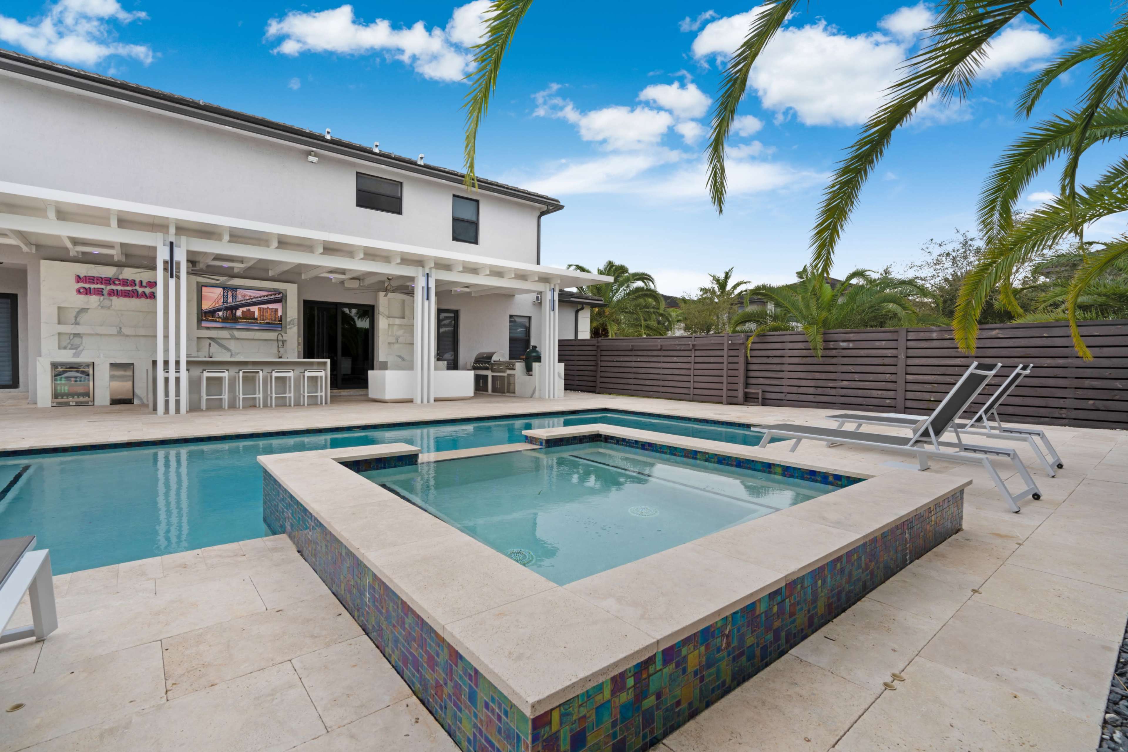 A modern outdoor area features a rectangular swimming pool surrounded by stone tiles, lounge chairs, and palm trees, with a covered patio in the background.