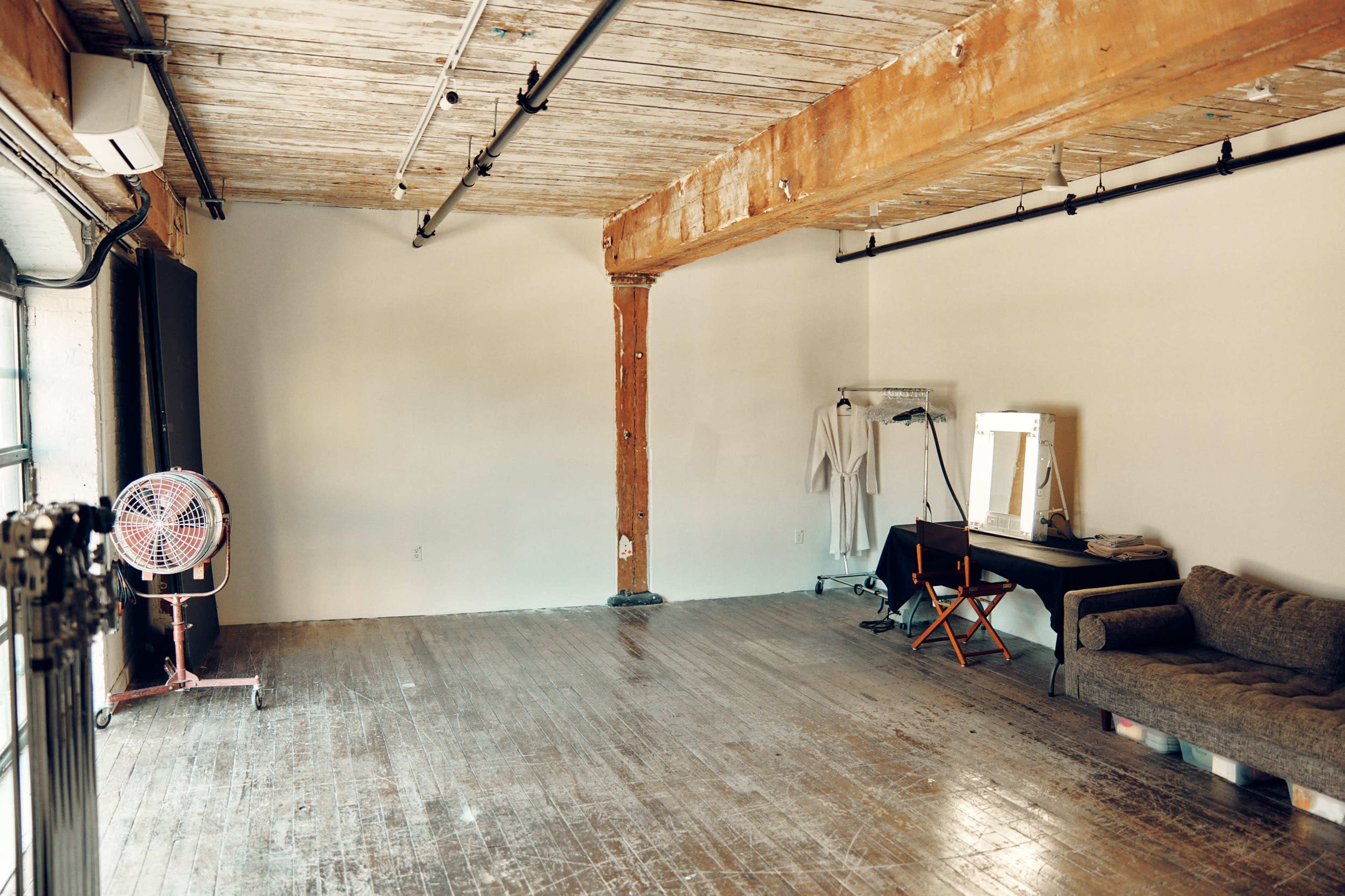 The image shows a spacious, unfinished studio with wooden floors and exposed beams, featuring a fan, a clothing rack, a bright mirror, and a couch.
