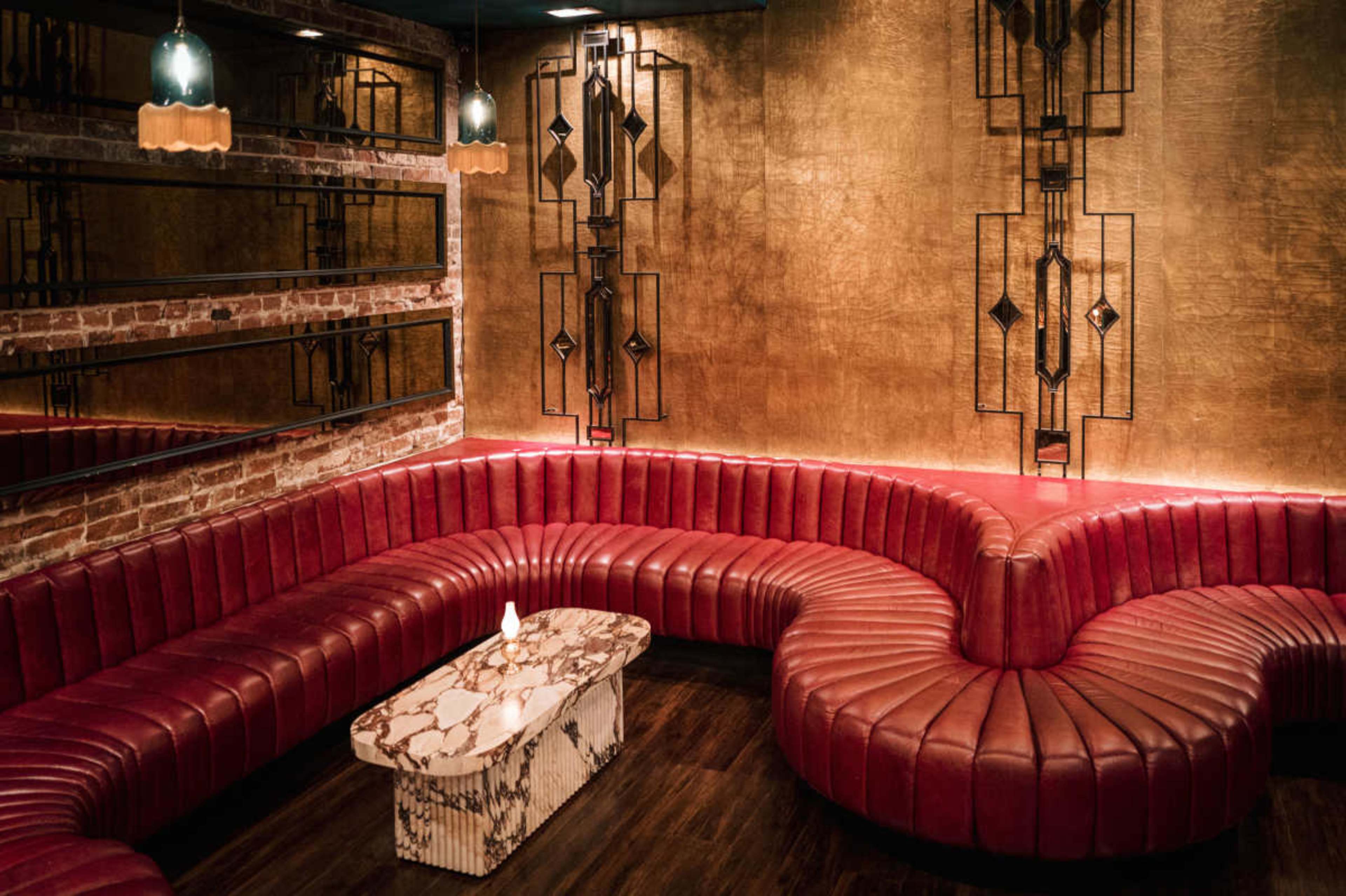 The image depicts a stylish lounge area with a large, curved red leather couch and a marble coffee table set against a backdrop of gold-colored walls and mirrors.