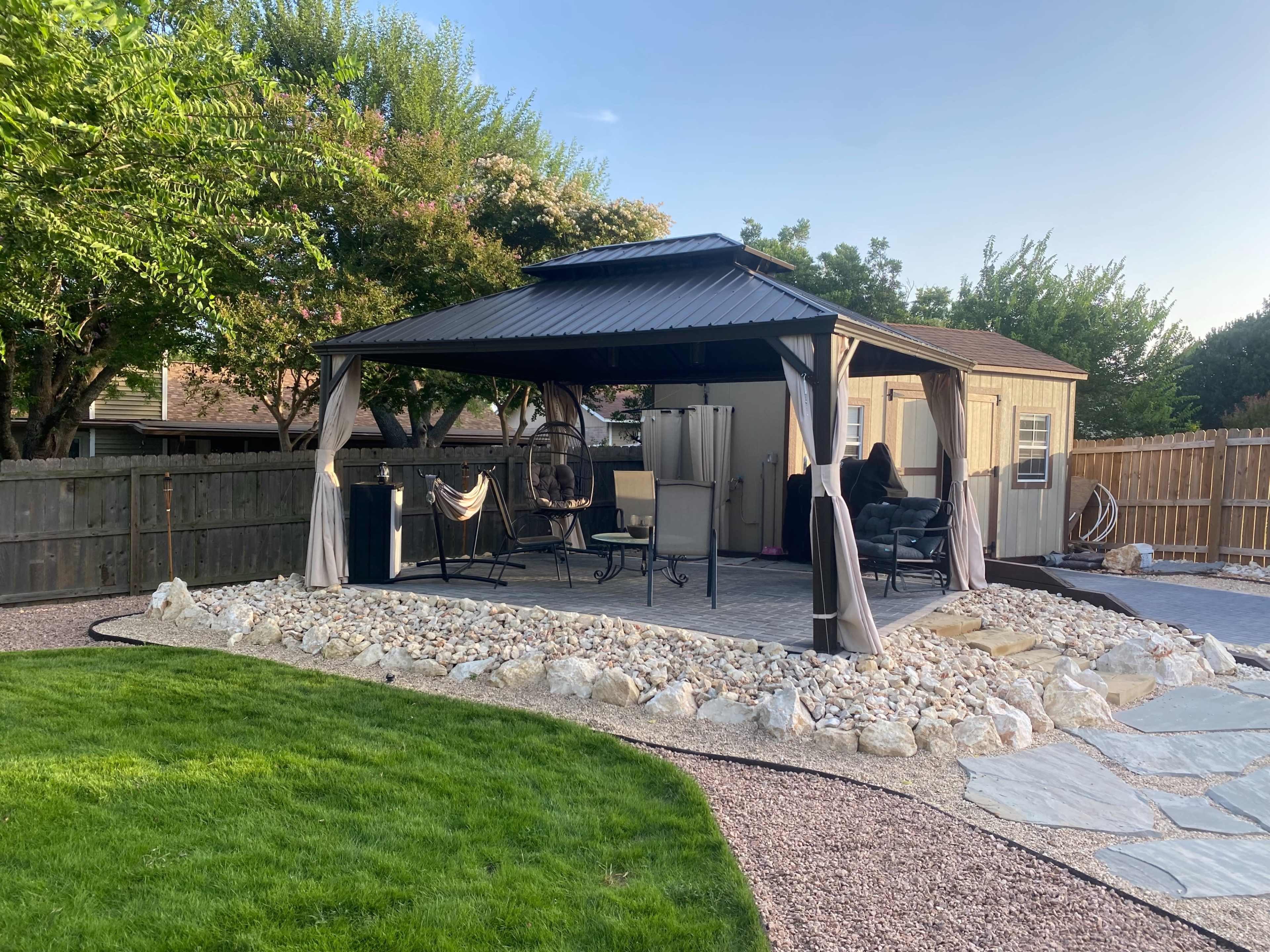 A gazebo with a metal roof sits on a stone patio surrounded by a grassy lawn and landscaped shrubs in a backyard.