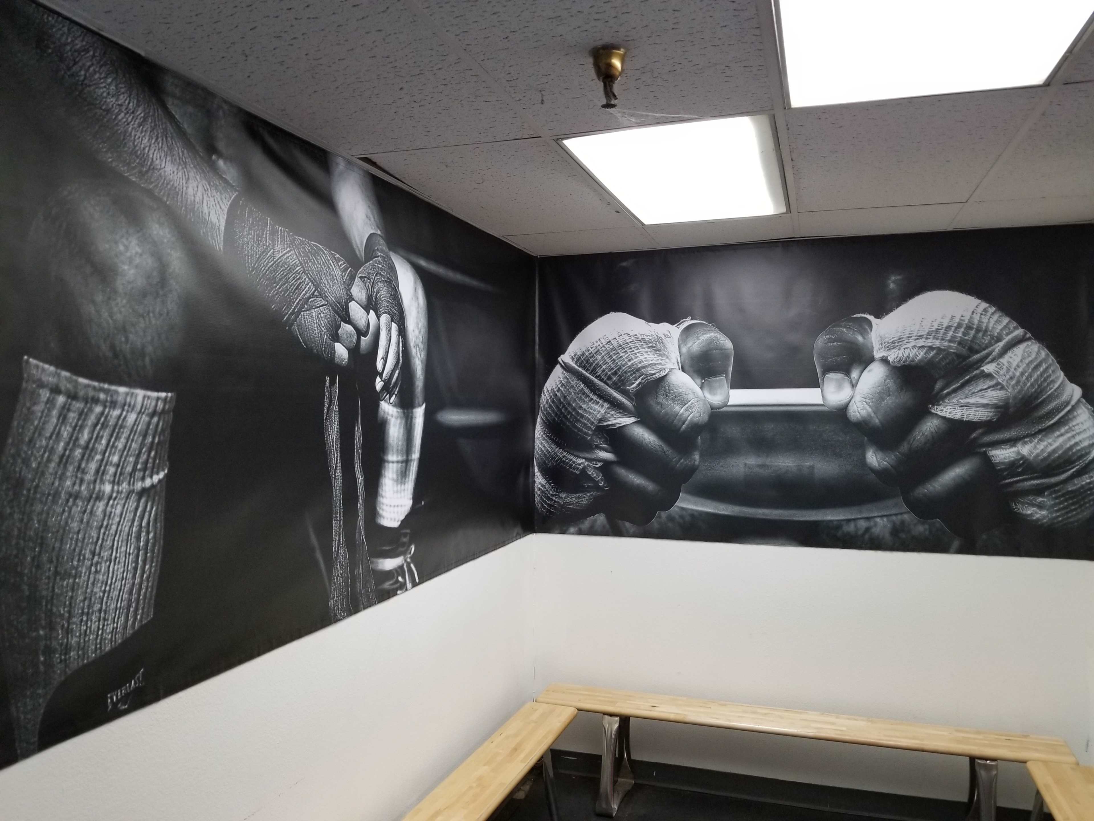The image shows a fitness studio corner with large black-and-white wall graphics depicting hands wrapped in gym tape gripping a barbell and a person preparing for a workout.