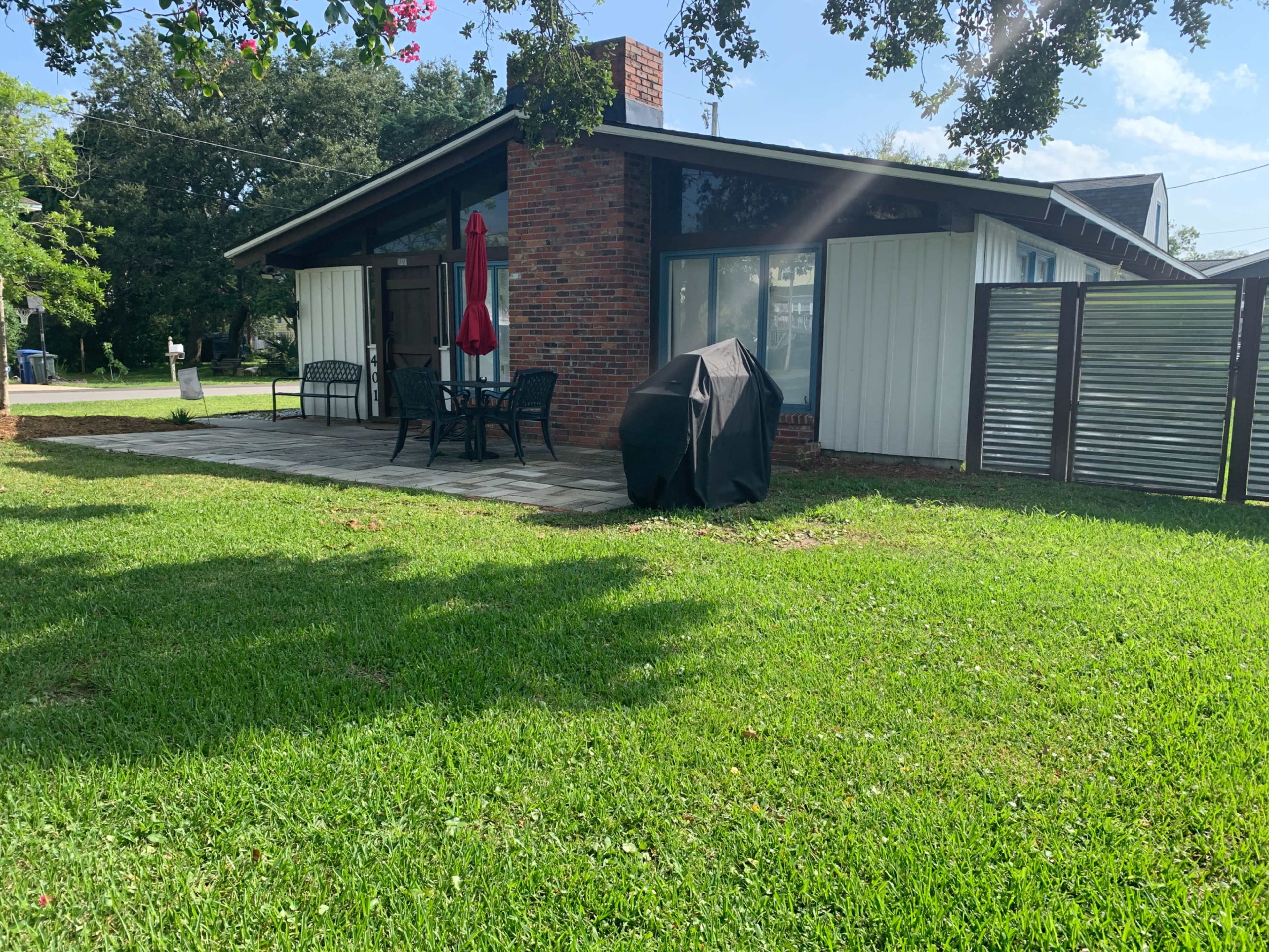 A single-story house with a red umbrella over a patio table, a charcoal grill, and grass surrounding it.