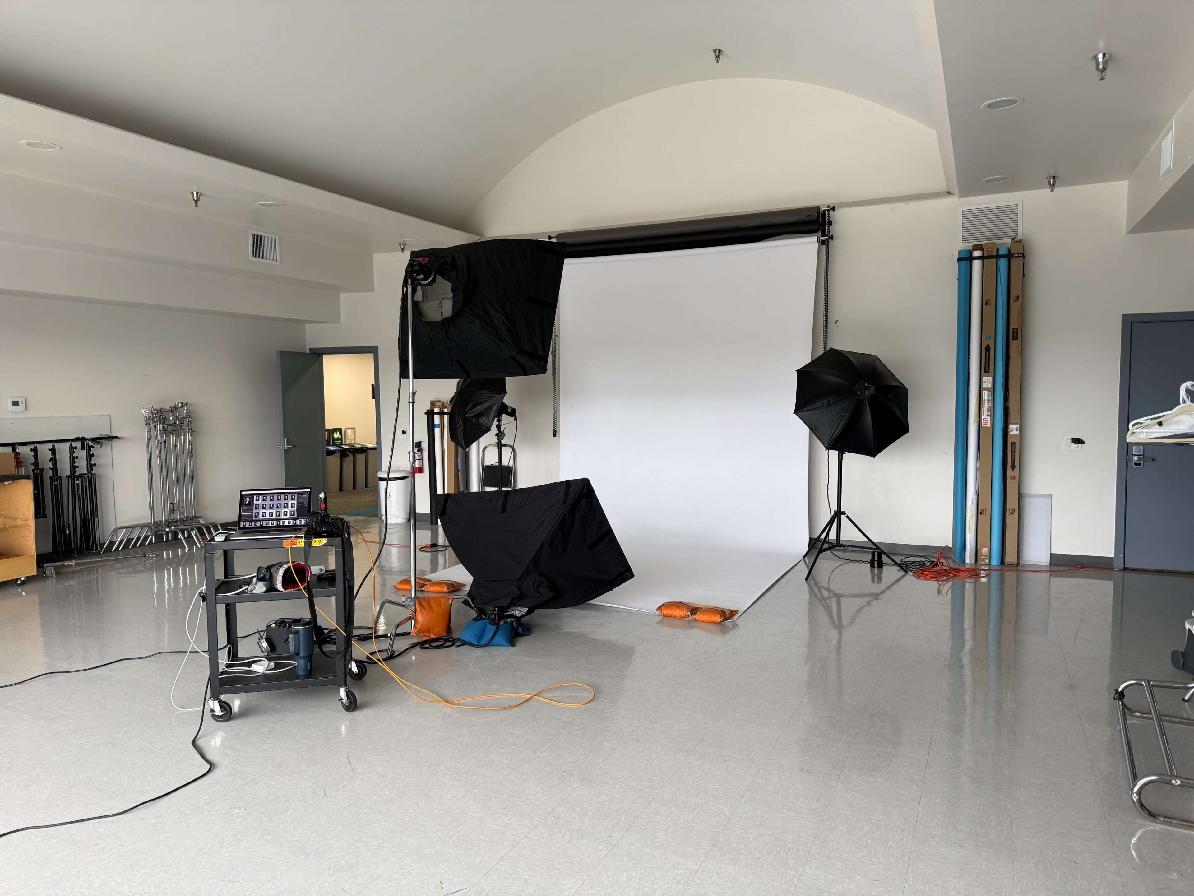 The image shows a photography studio setup with a white backdrop, softboxes, and lighting equipment arranged around a clean, open space.