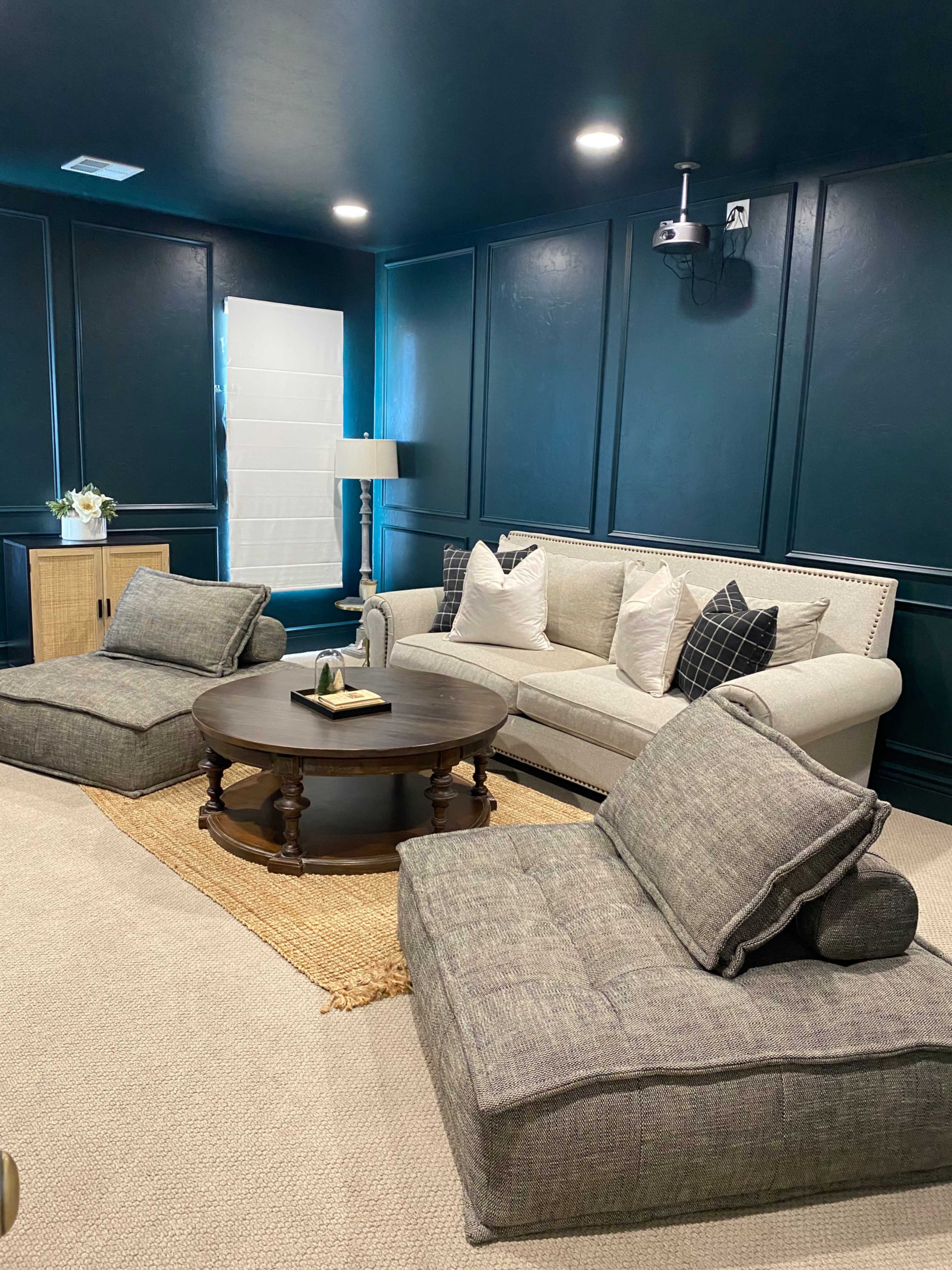 A cozy living room with dark blue walls, a light-colored sofa, two oversized floor cushions, a round coffee table, and a textured rug.