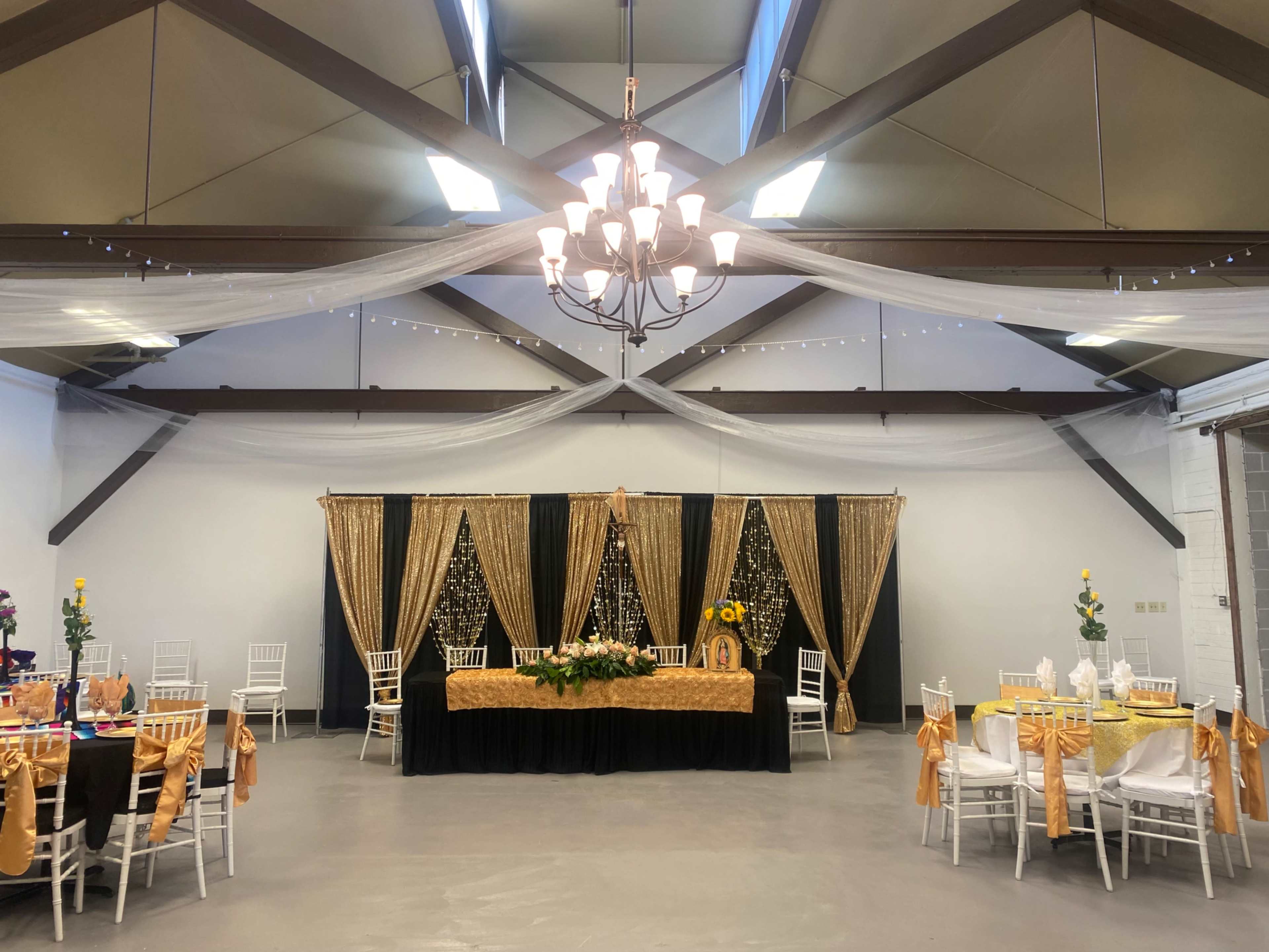 A spacious event hall features elegantly arranged tables with gold and white decorations, a large black table at the front, and draped fabric from the ceiling.